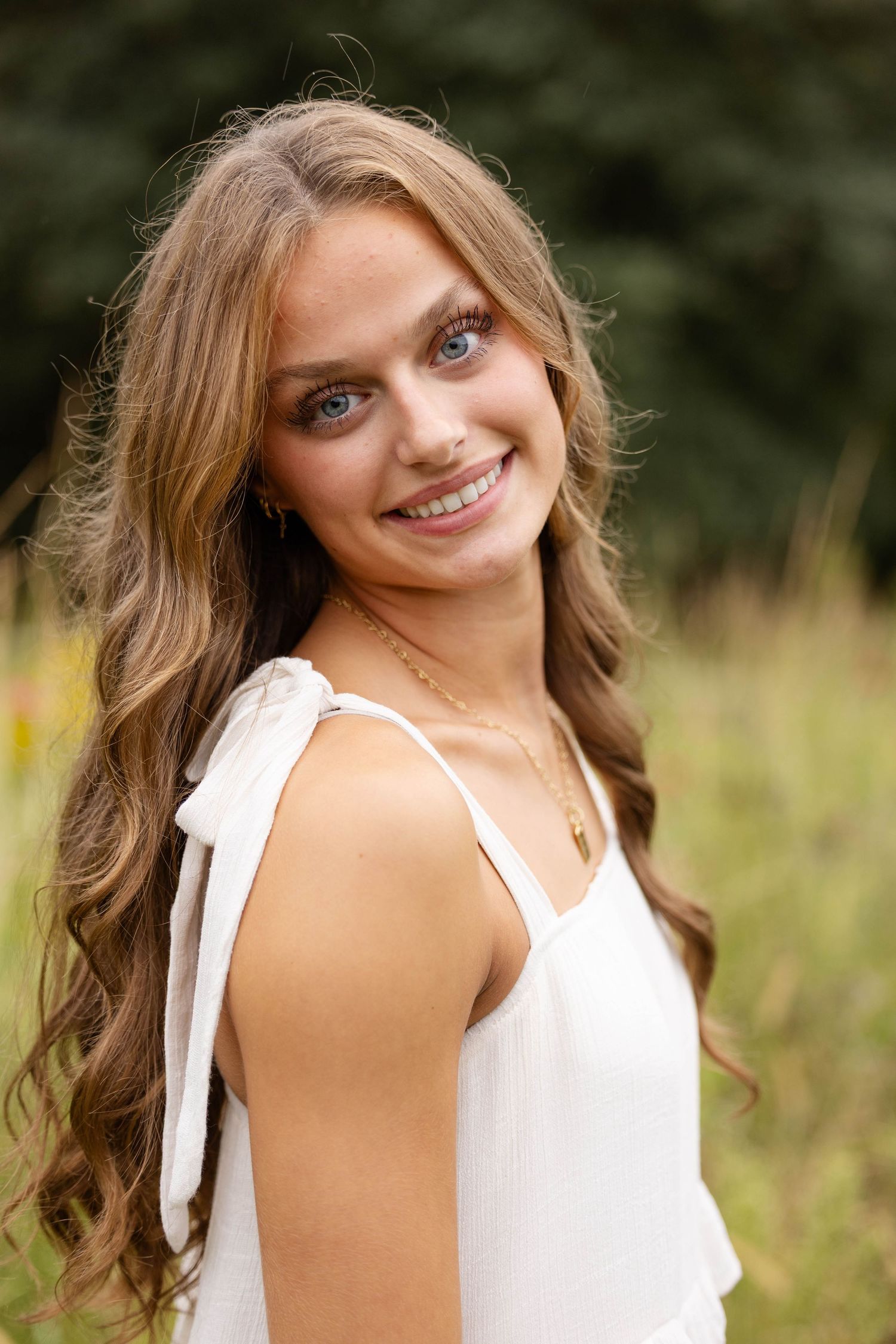 Smiling young woman with long wavy light brown hair wearing a white tie-shoulder top and gold necklace, posing in a sunlit grassy field.