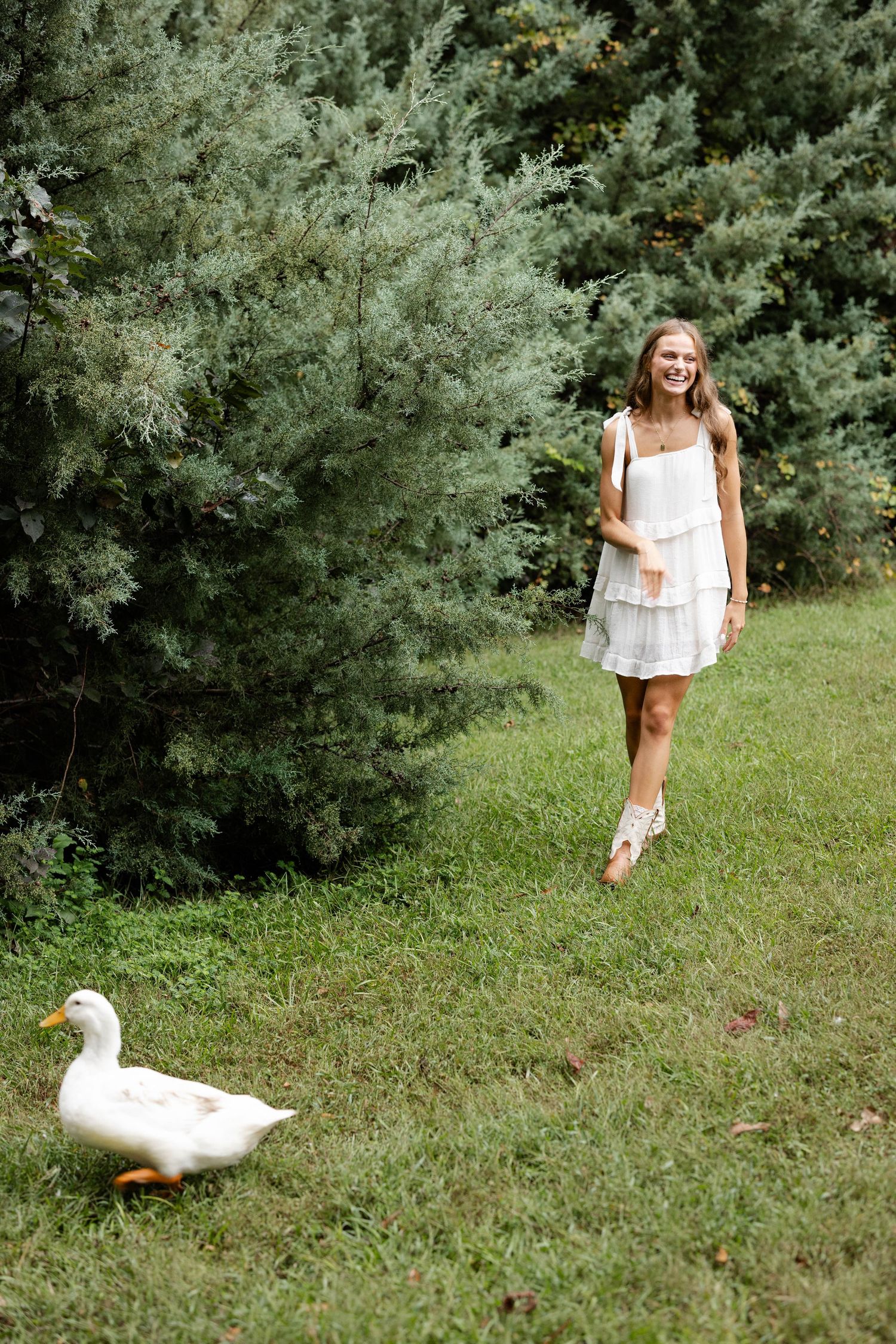 Smiling young woman in a white sundress walks barefoot across a green lawn toward a white duck near tall evergreen trees.