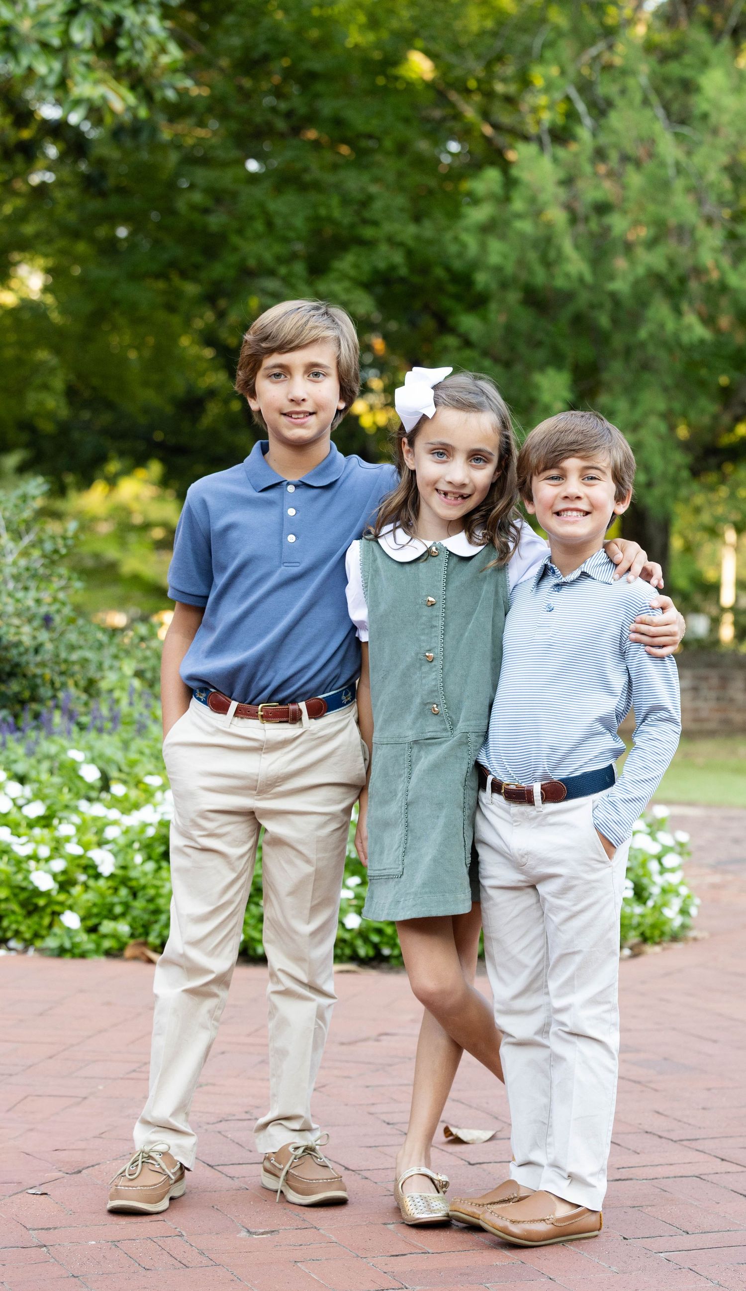Three smiling children in coordinated preppy outfits pose arm in arm on a brick path in front of green trees and white flowers.