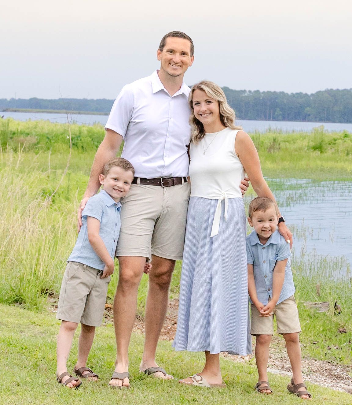 Family of four posing outdoors near a lake, parents with two young boys in coordinating outfits on a sunny day.