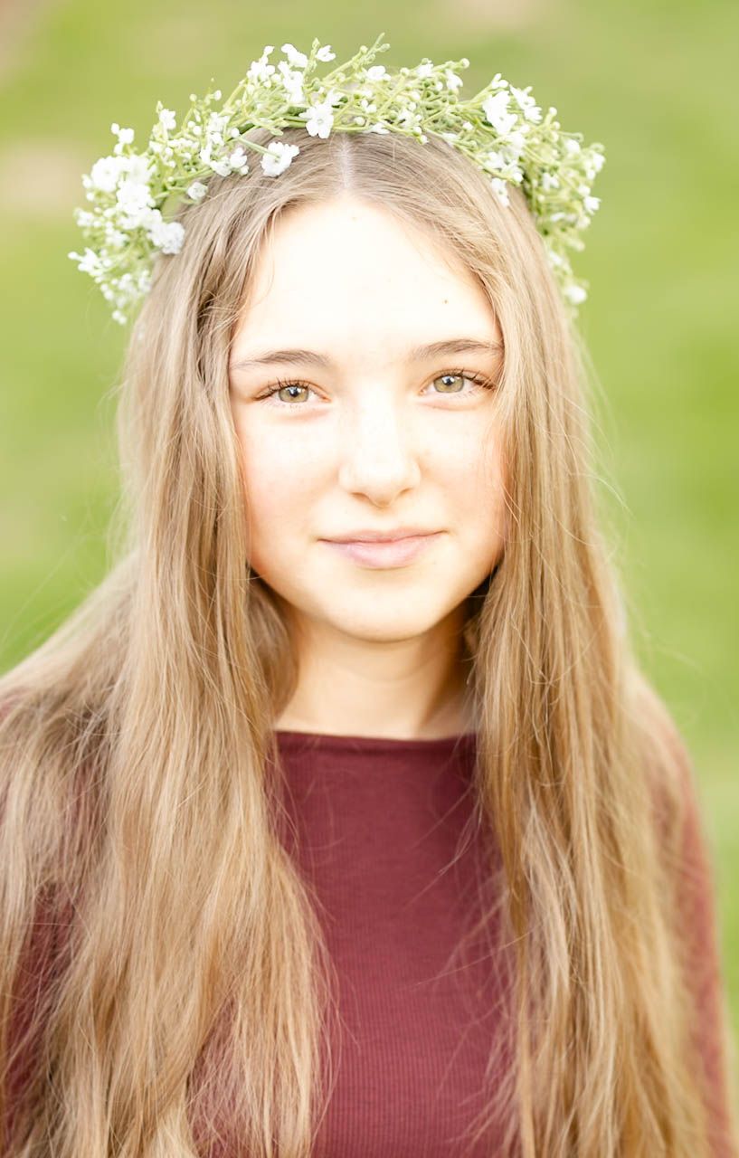 A teenage girl with long blonde hair wears a white flower crown, smiling softly outdoors.