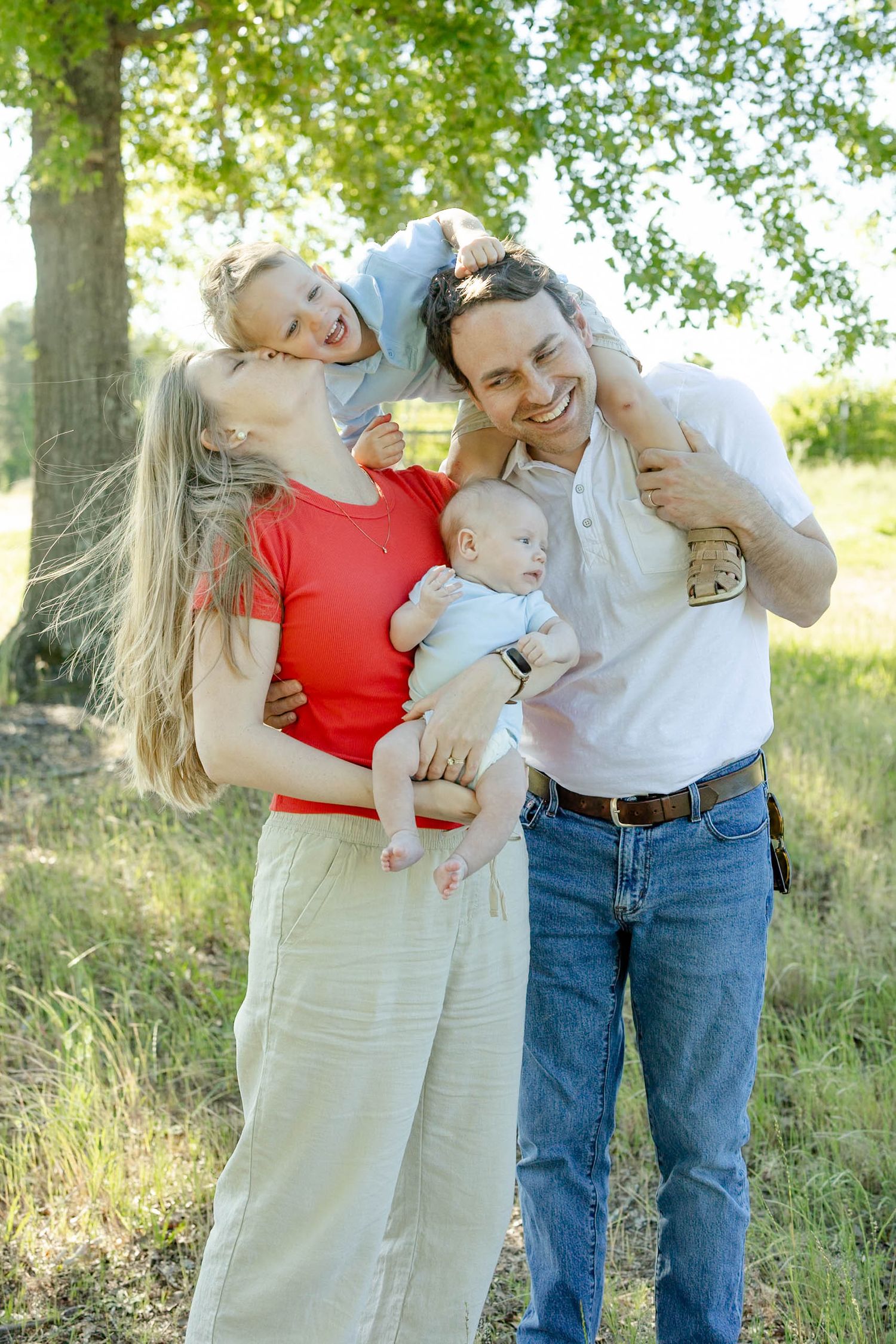 Happy family of four posing outdoors, parents holding baby and toddler, smiling in a green park setting.
