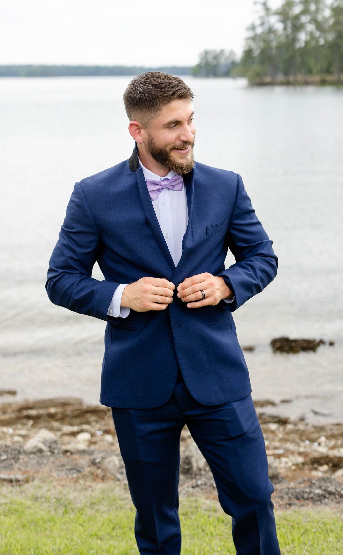 A smiling groom in a navy blue suit and lavender bow tie stands outdoors near a lake on his wedding day.