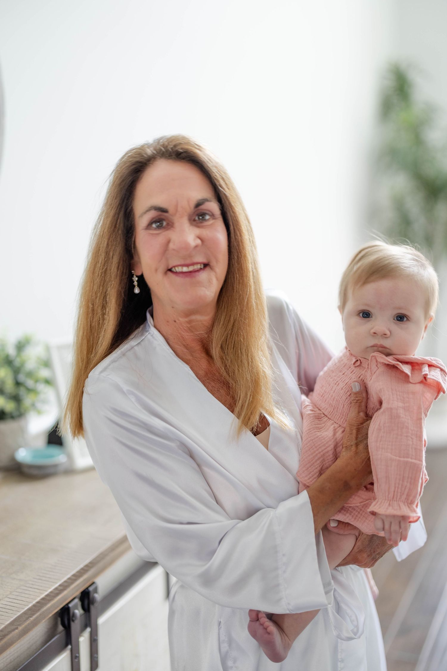 A smiling grandmother in a white robe holding an adorable baby dressed in pink indoors.