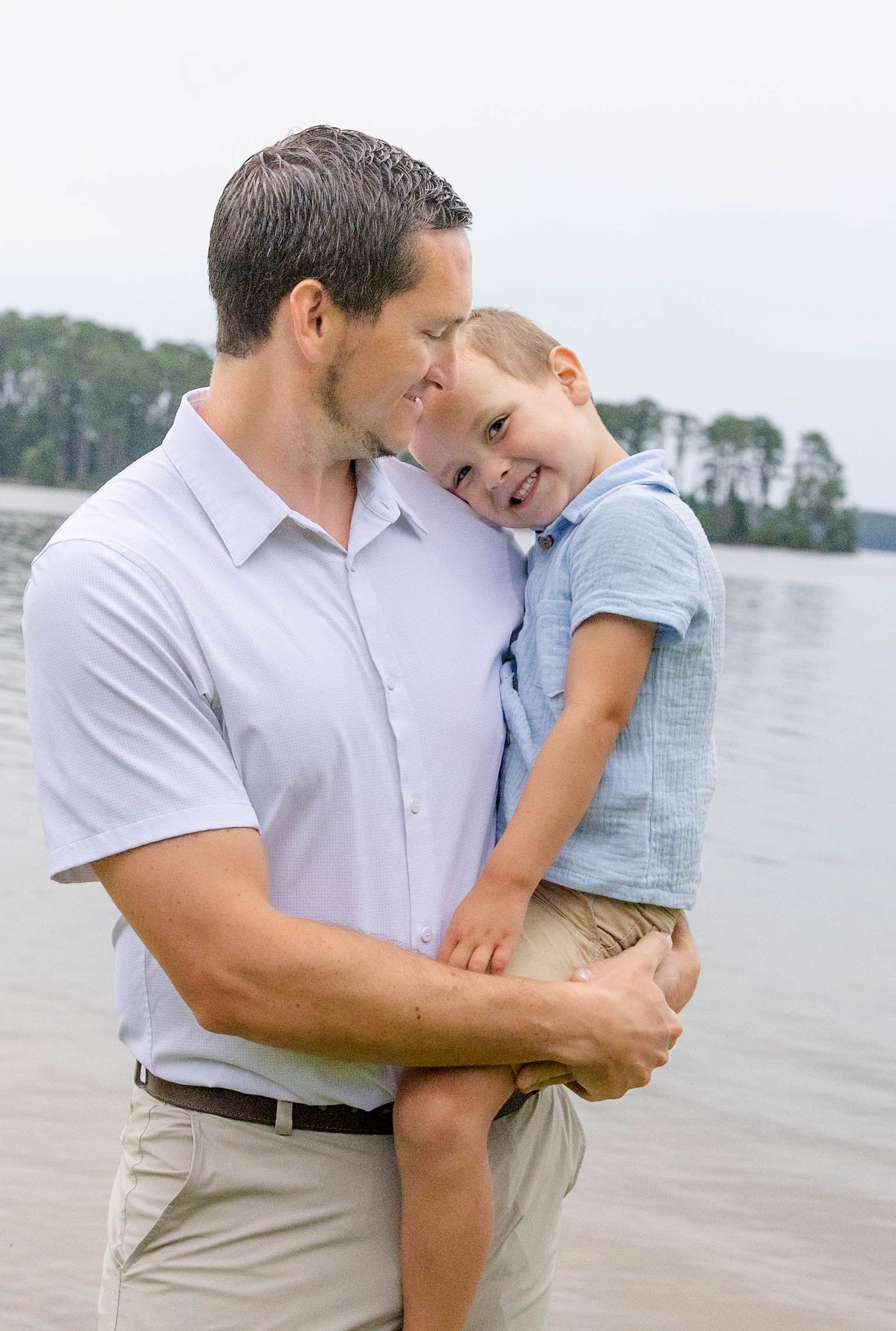 A father lovingly holds his smiling young son near a lake, both wearing light blue shirts outdoors.