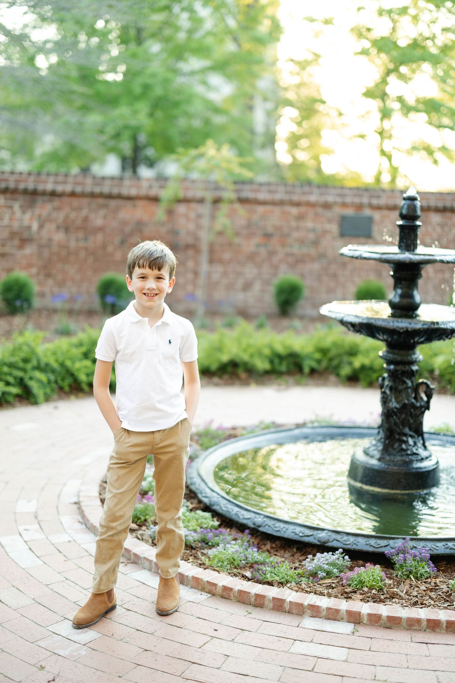 A smiling boy in white polo and khaki pants stands confidently near an ornate fountain in a brick garden.