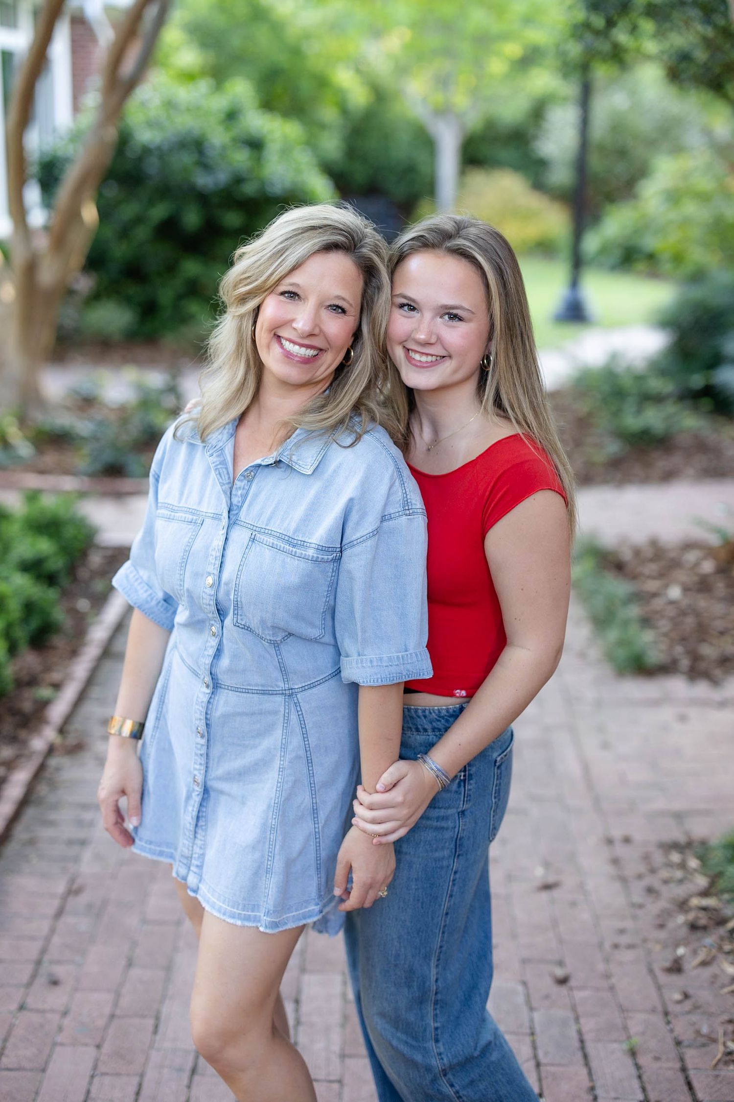 Smiling mother and teenage daughter posing together on a brick path surrounded by lush greenery.