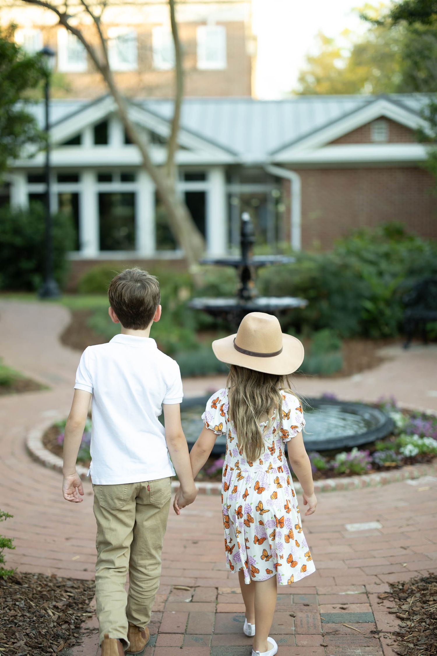 Two children holding hands walking toward a fountain on a brick pathway outside a building.