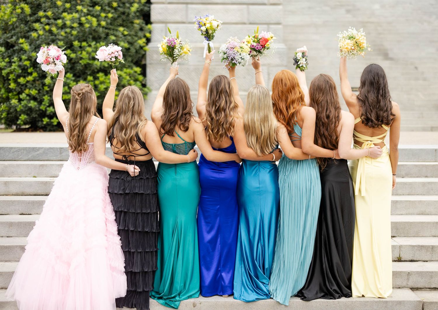 Bridesmaids in colorful gowns raise bouquets on steps, viewed from behind, with bride in pink ruffled dress.