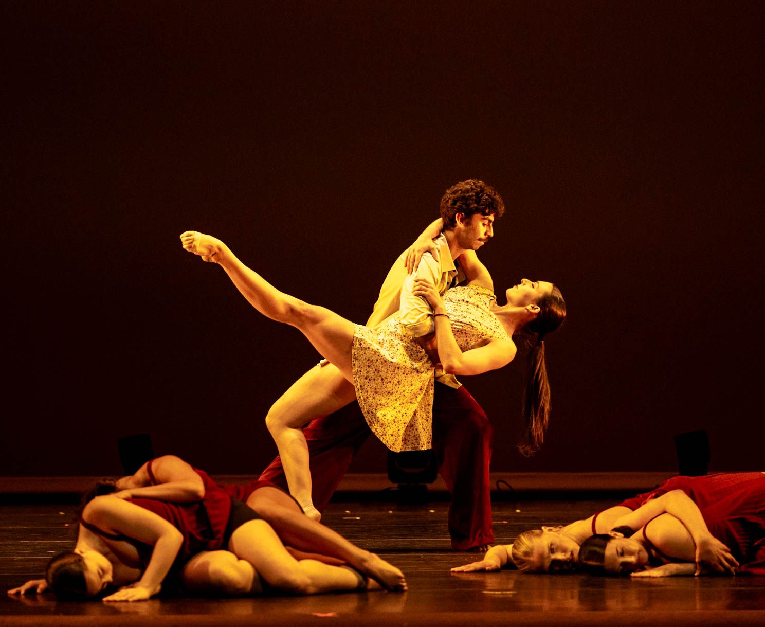 Two ballet dancers perform a dramatic lift on a dimly lit stage while other dancers in red costumes lie curled on the floor.