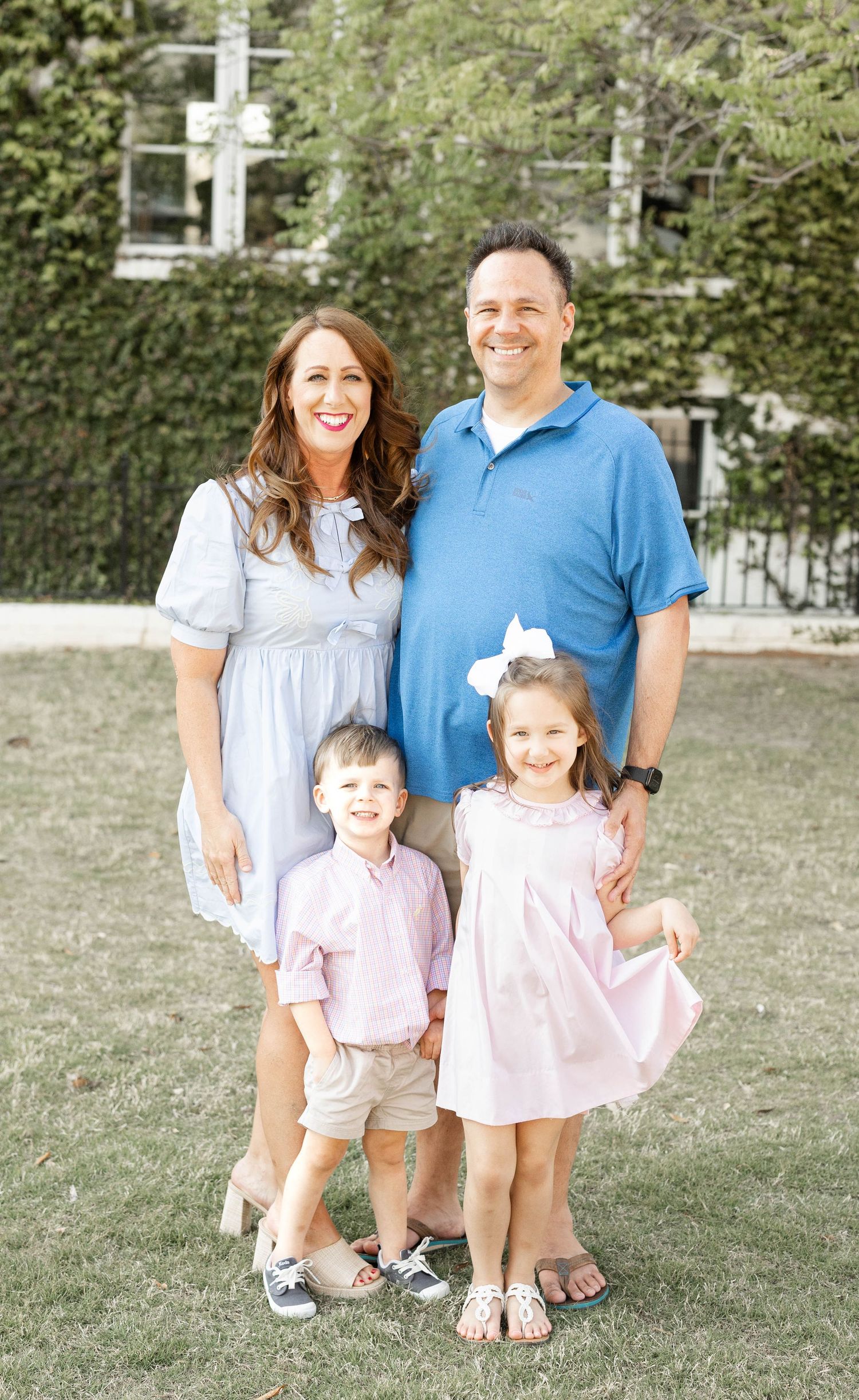 Happy family of four posing outdoors on green grass with two young children dressed in pink.