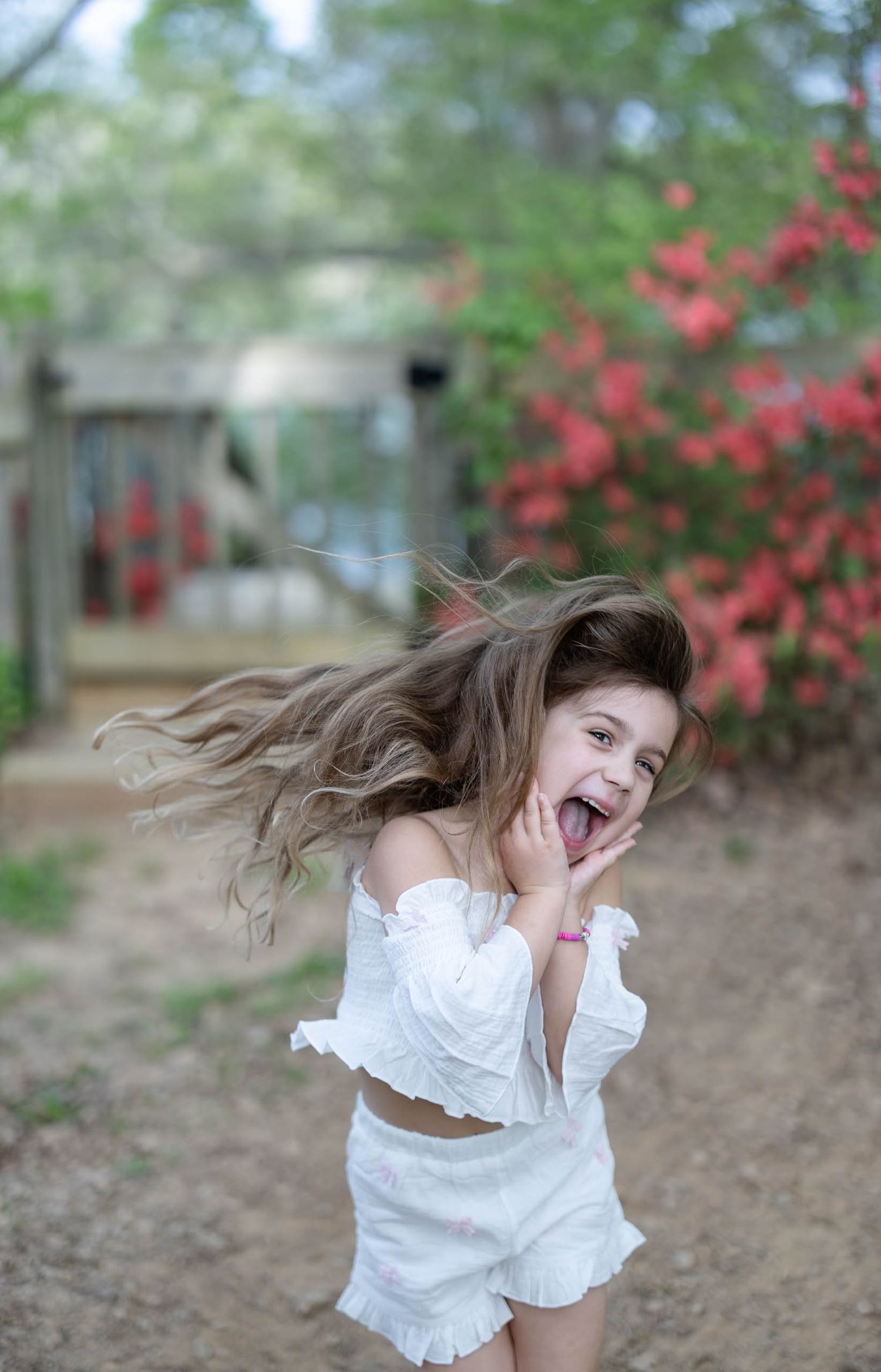 A gleeful little girl in white outfit spins with mouth open wide as her long hair flies in an outdoor setting.
