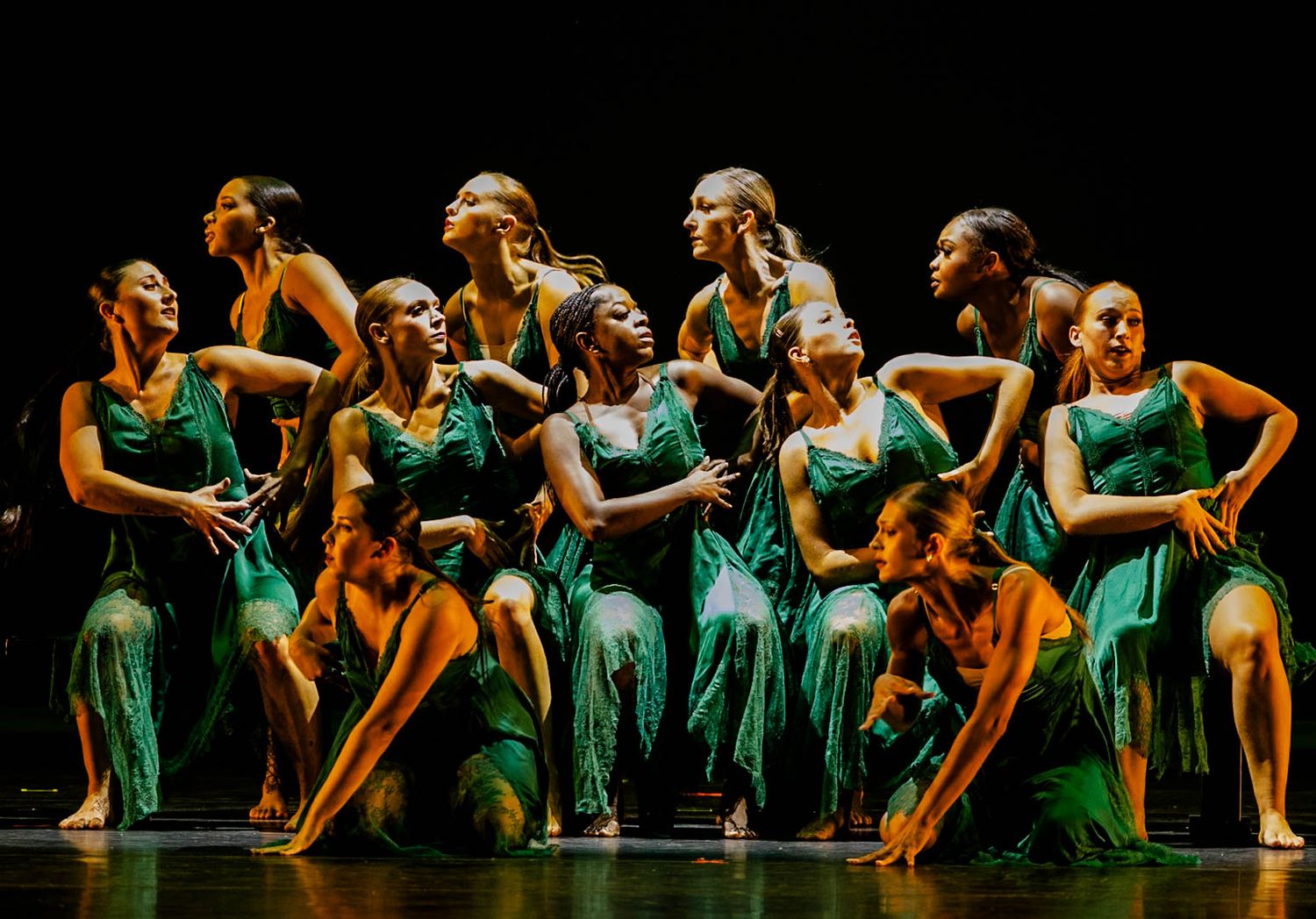 Group of dancers in flowing green dresses pose in an expressive contemporary dance formation on a dark stage.