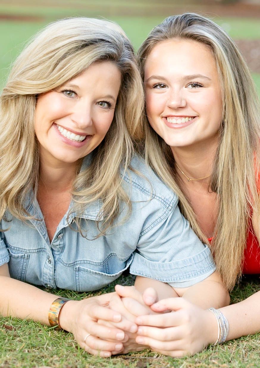 Two blonde women smile while lying on grass, wearing casual outfits in a sunny outdoor portrait.