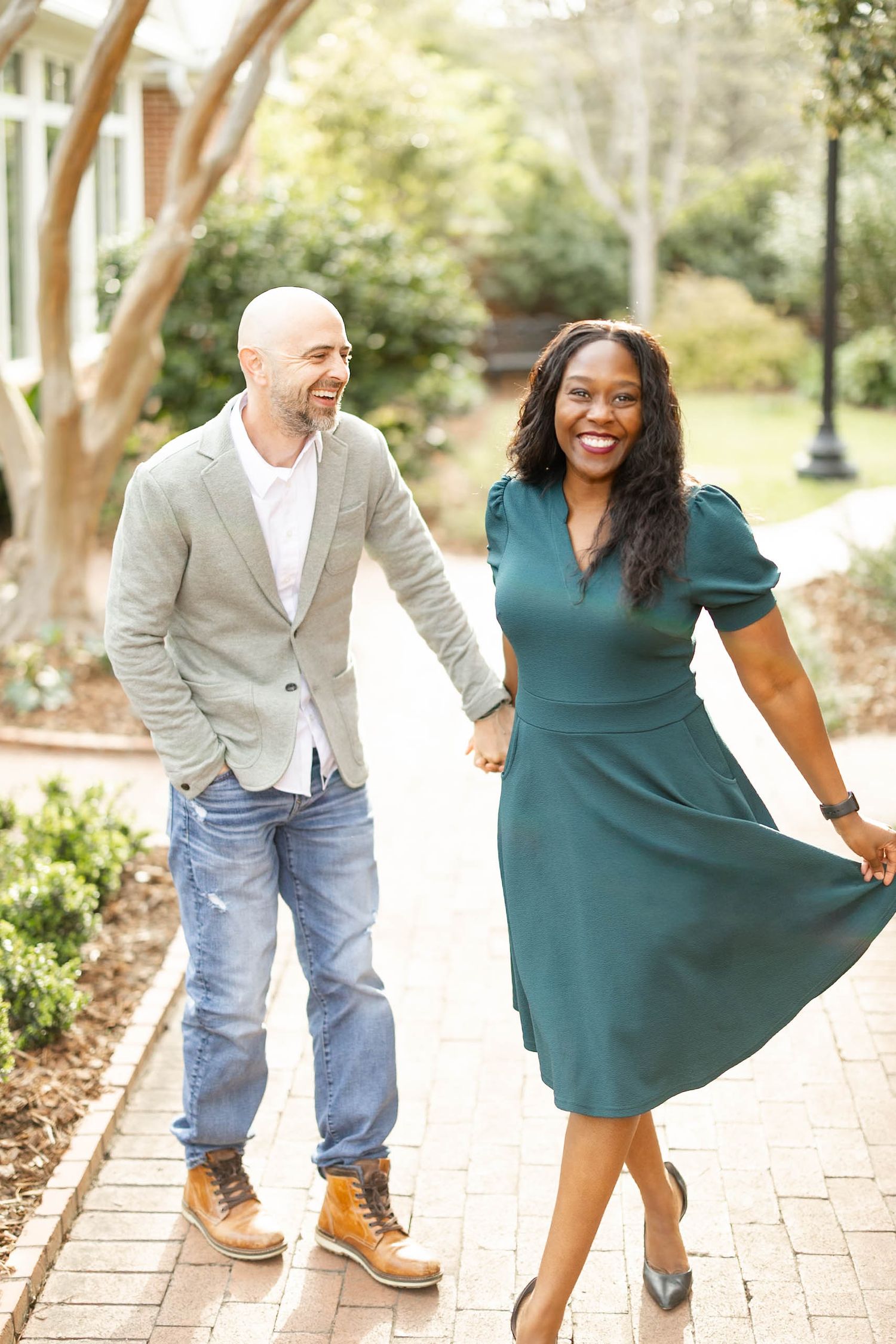 A joyful interracial couple walks hand in hand on a brick path, laughing in a garden setting.