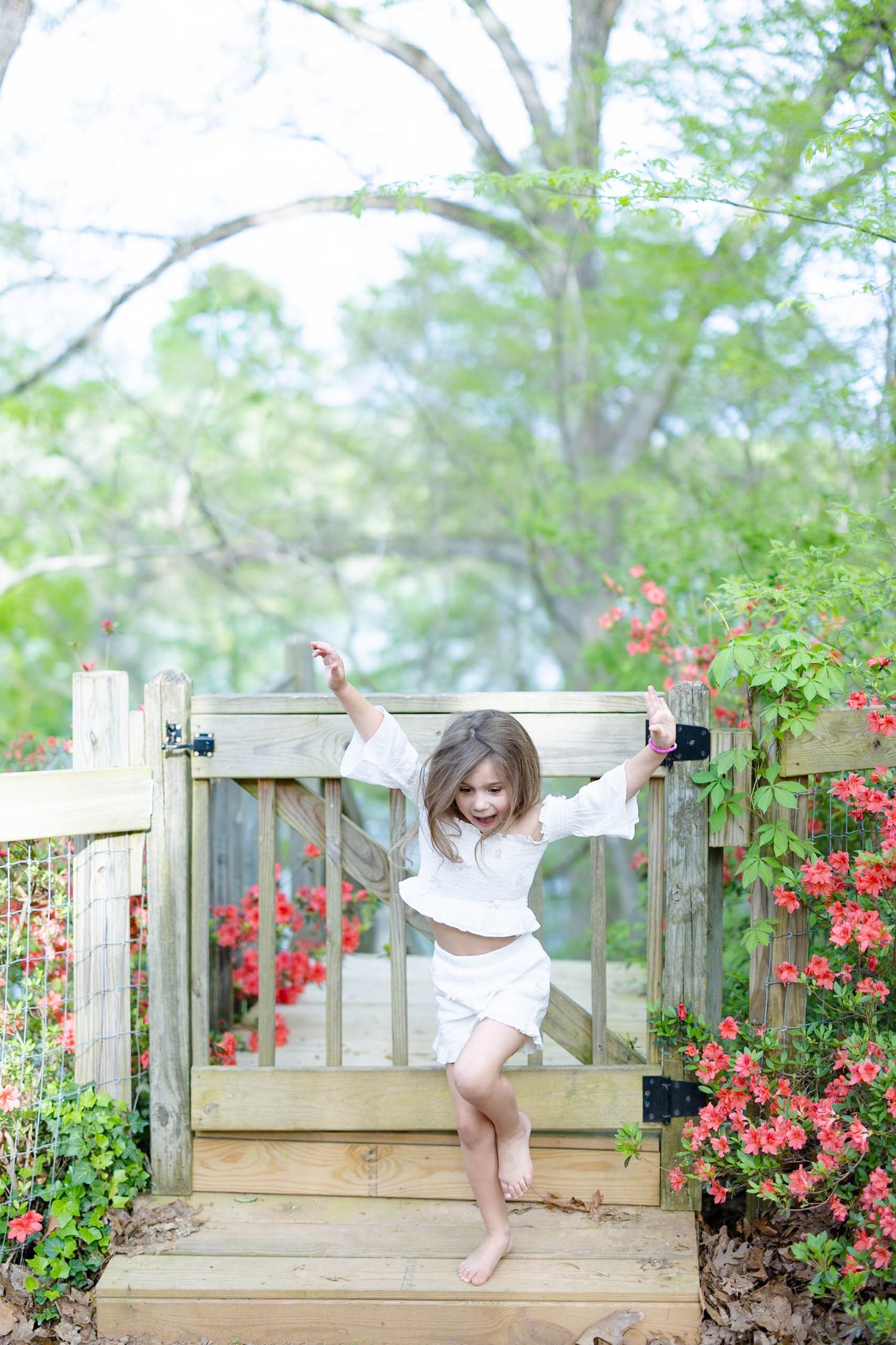 Happy barefoot girl in white outfit playing on a wooden garden gate surrounded by red flowers.