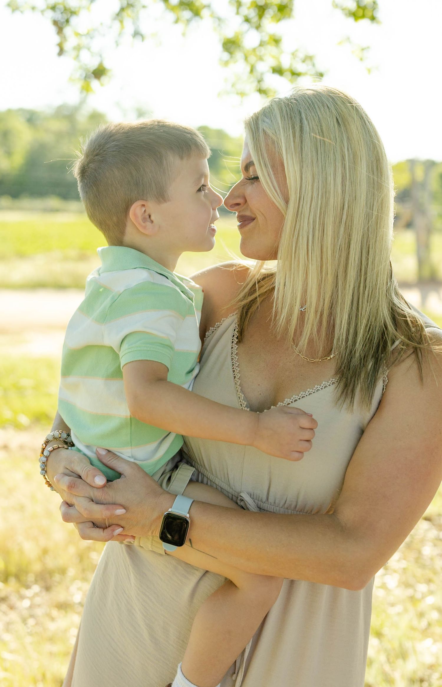 A blonde woman holds and smiles nose-to-nose with a young boy in a green striped shirt outdoors.