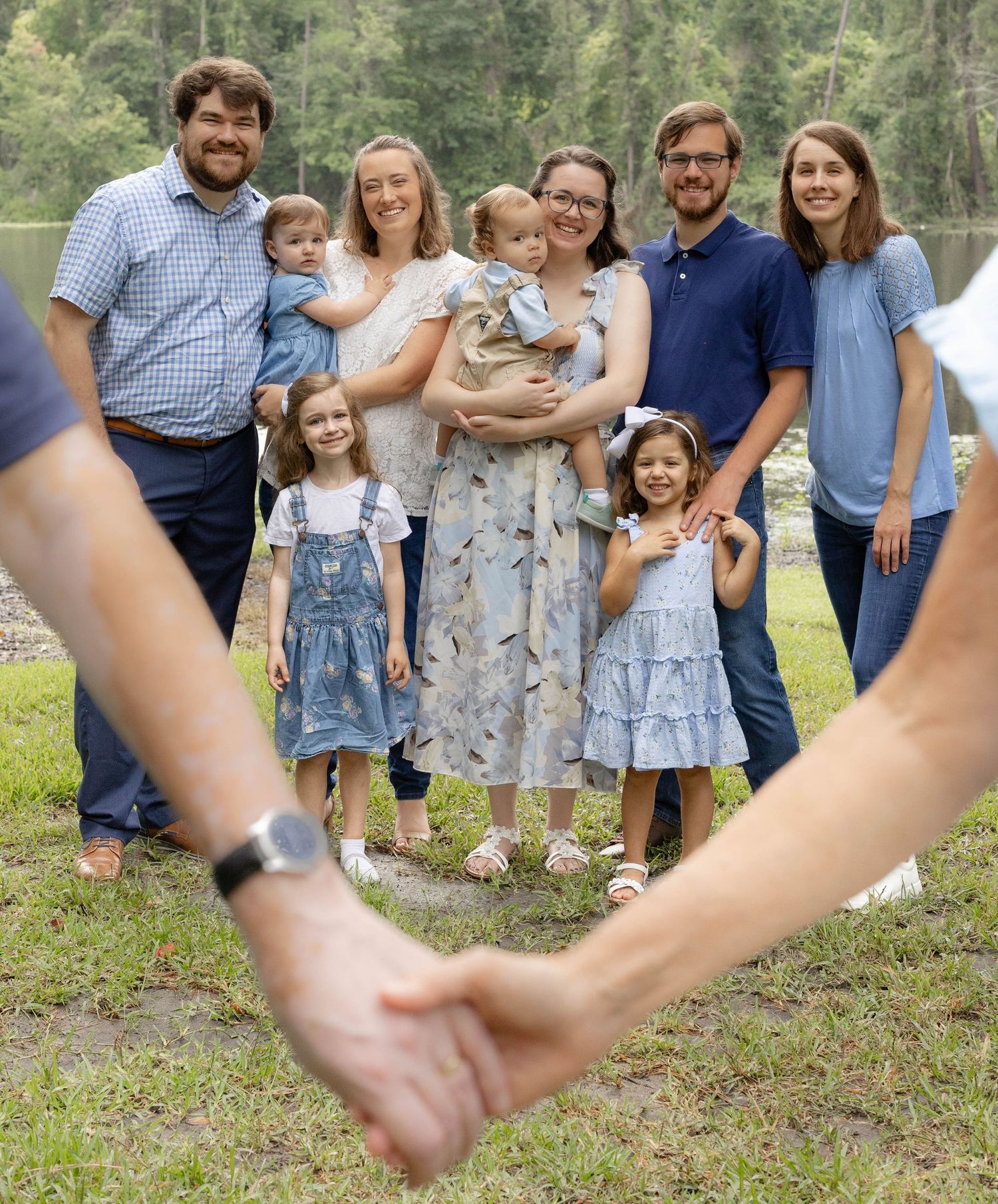 Smiling family of eight with young children posing outdoors by a lake as two people in the foreground hold hands.