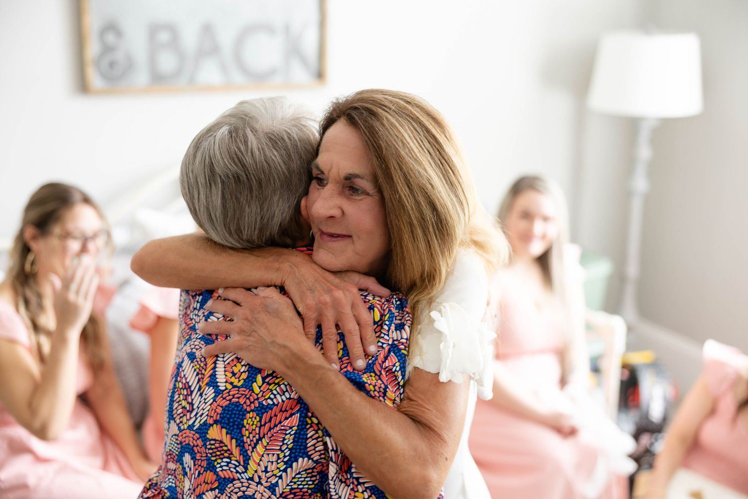 Two women share a warm embrace at a bridal shower with bridesmaids in pink in the background.