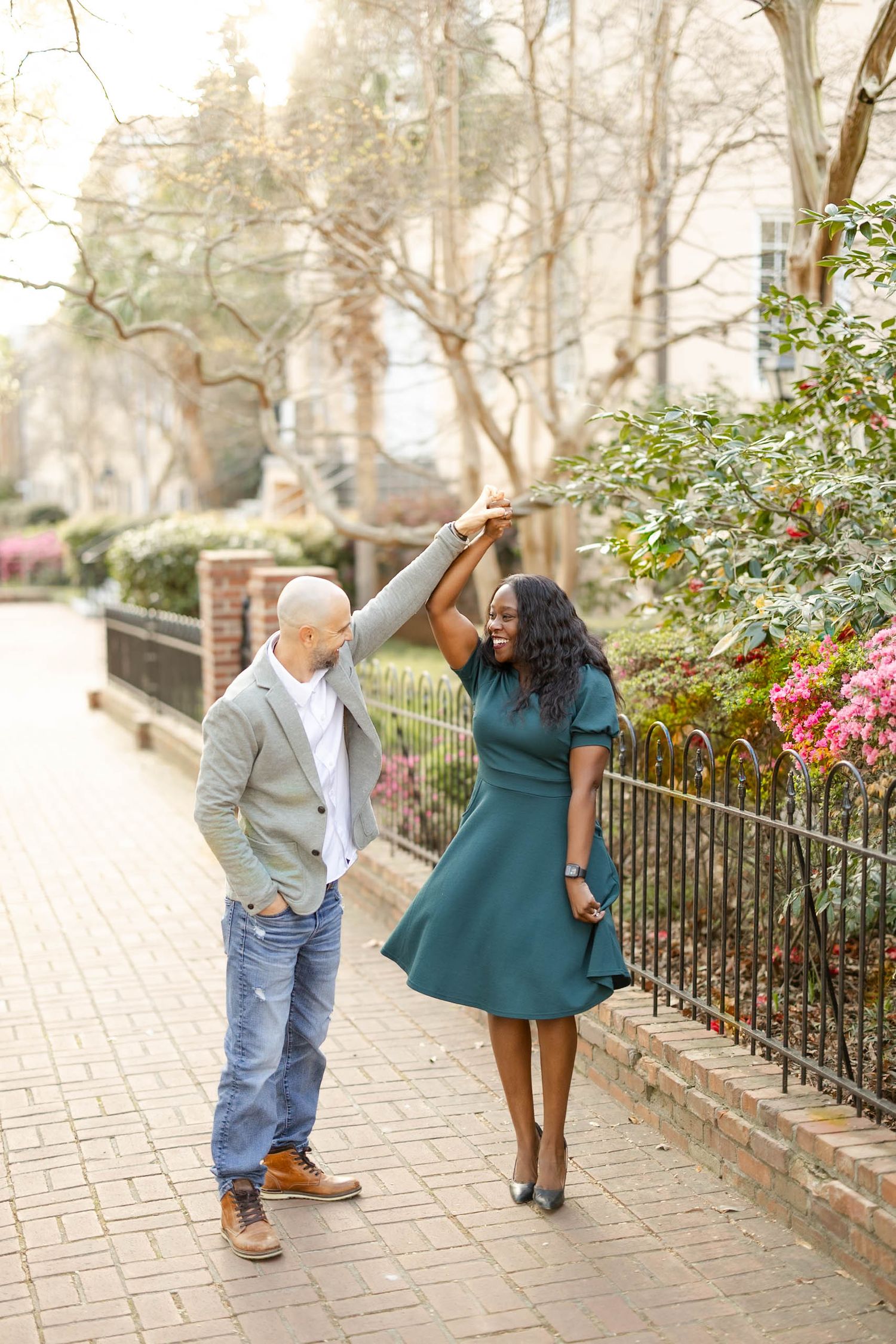 Joyful interracial couple dancing on a brick pathway surrounded by blooming flowers and greenery.