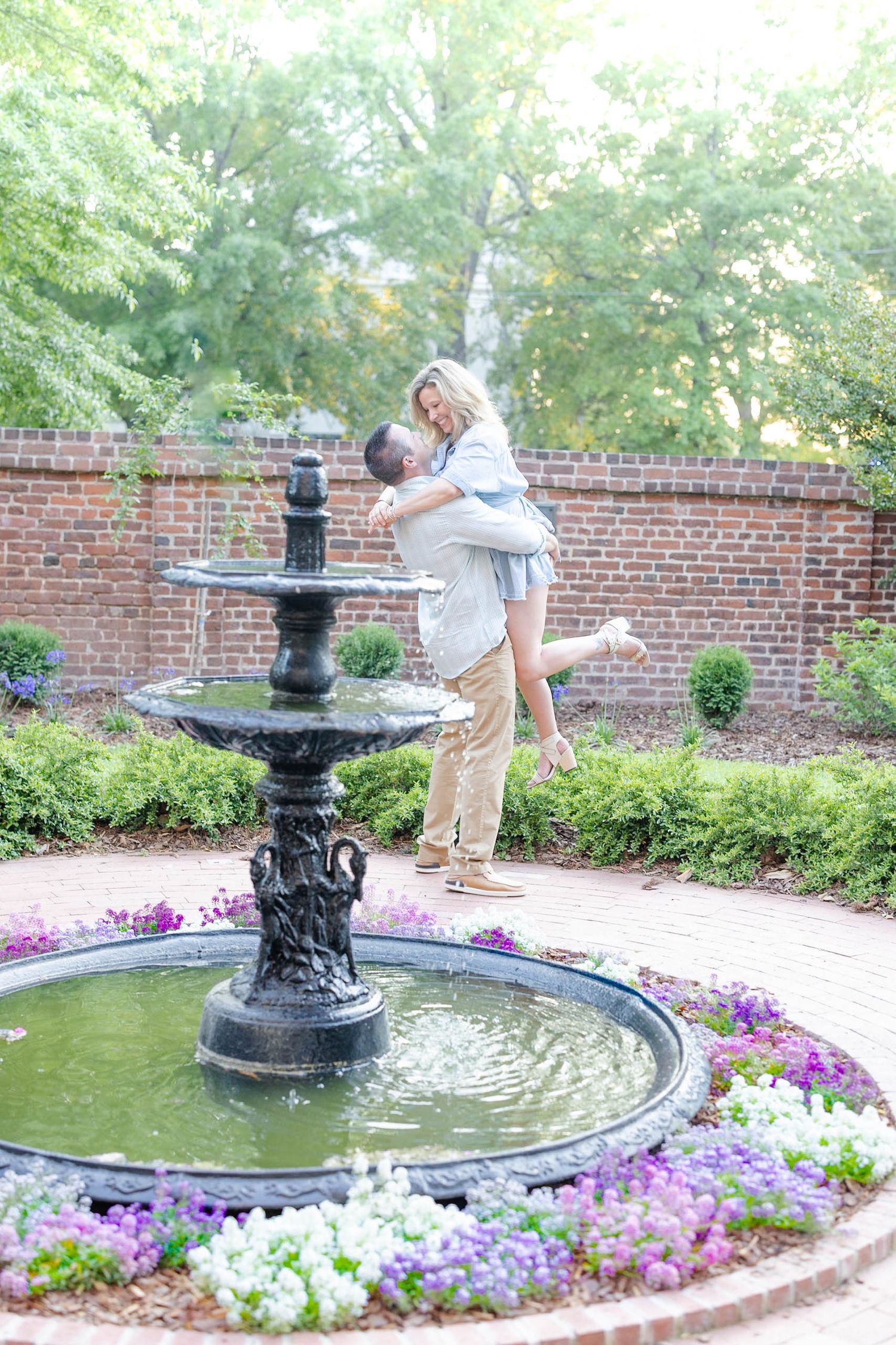 Man lifting smiling woman near a decorative garden fountain surrounded by colorful flowers.