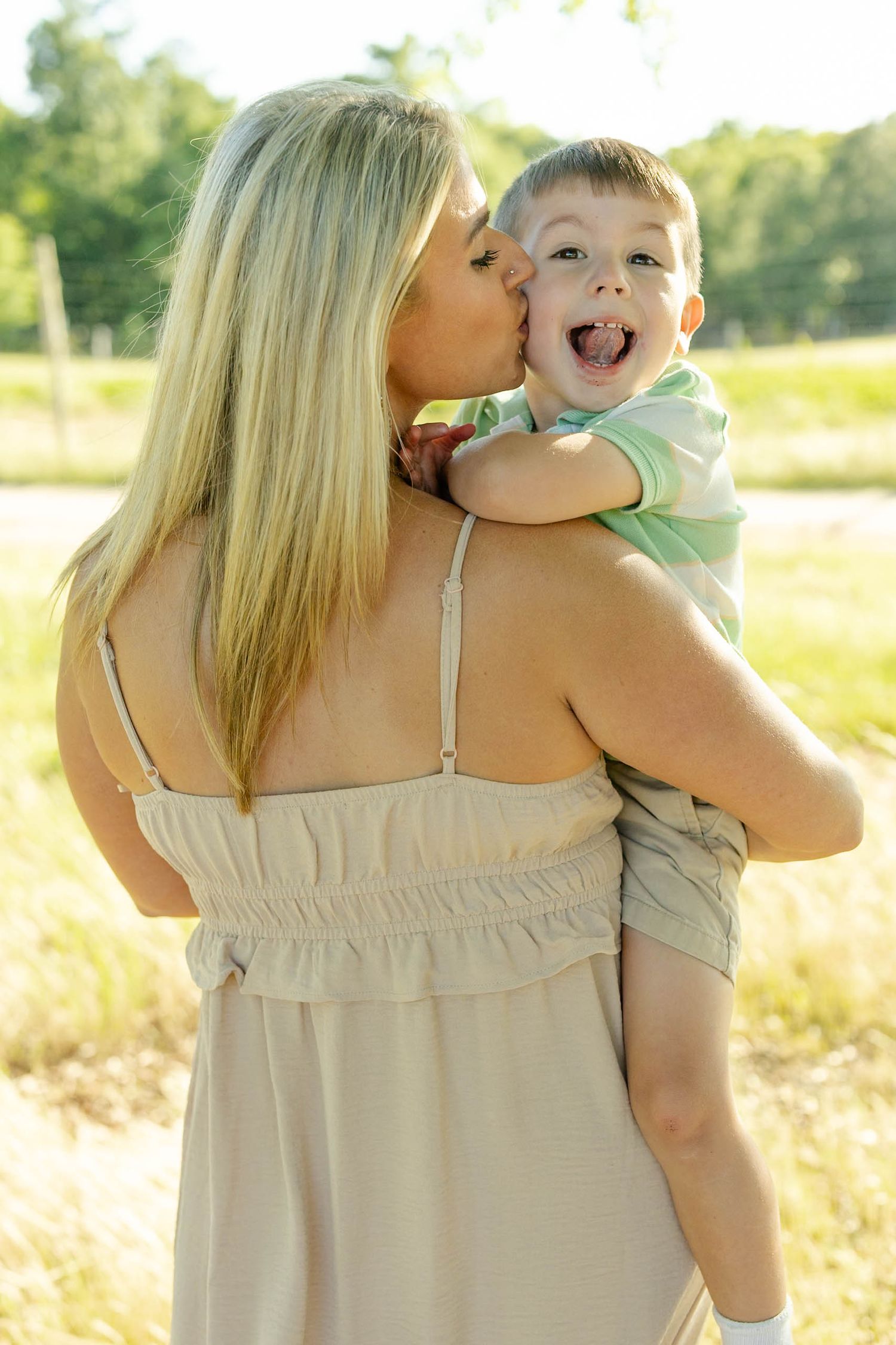 A blonde woman in a beige dress kisses a laughing young boy she holds in her arms outdoors.