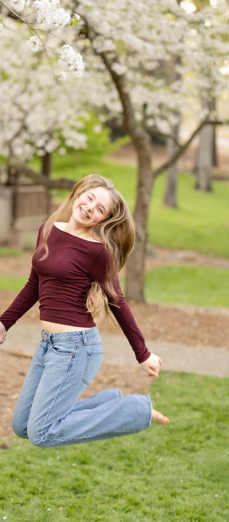 Joyful teenage girl jumping in the air in front of blooming white spring cherry trees.