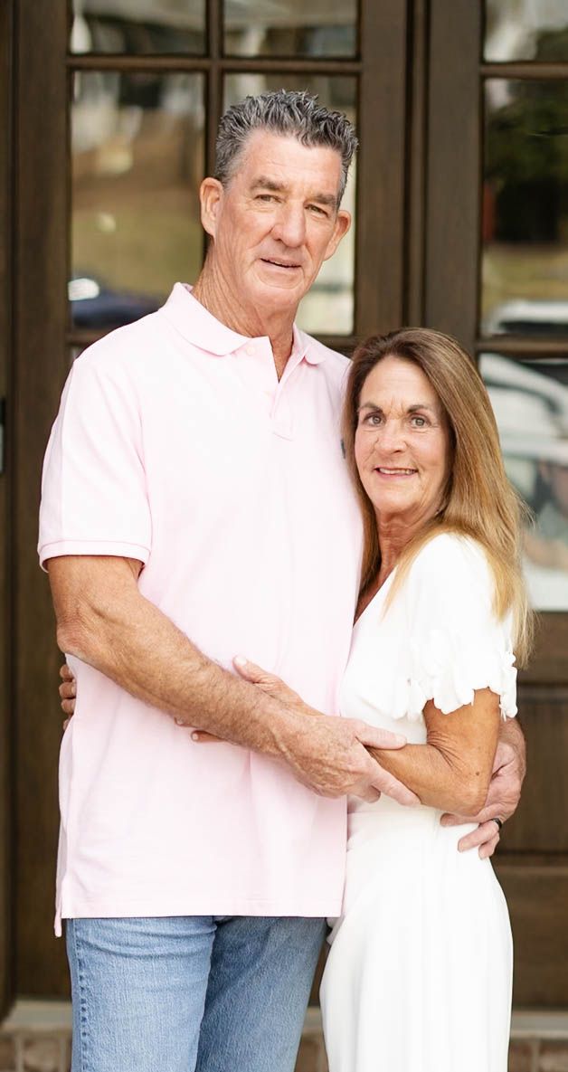 An older couple embraces and smiles outdoors, man in pink polo and woman in white dress, standing before glass doors.