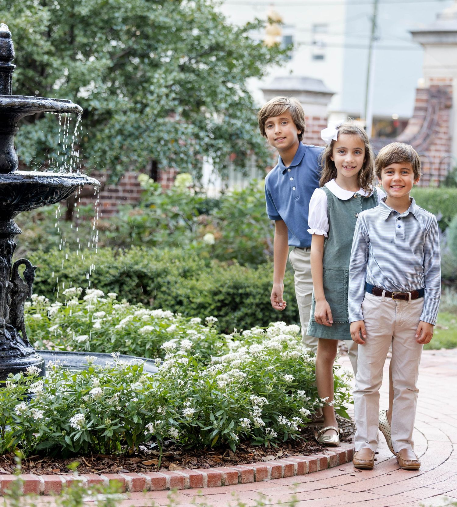 Three children in coordinated casual outfits stand on a brick garden path beside a tiered stone fountain and blooming white flowers.
