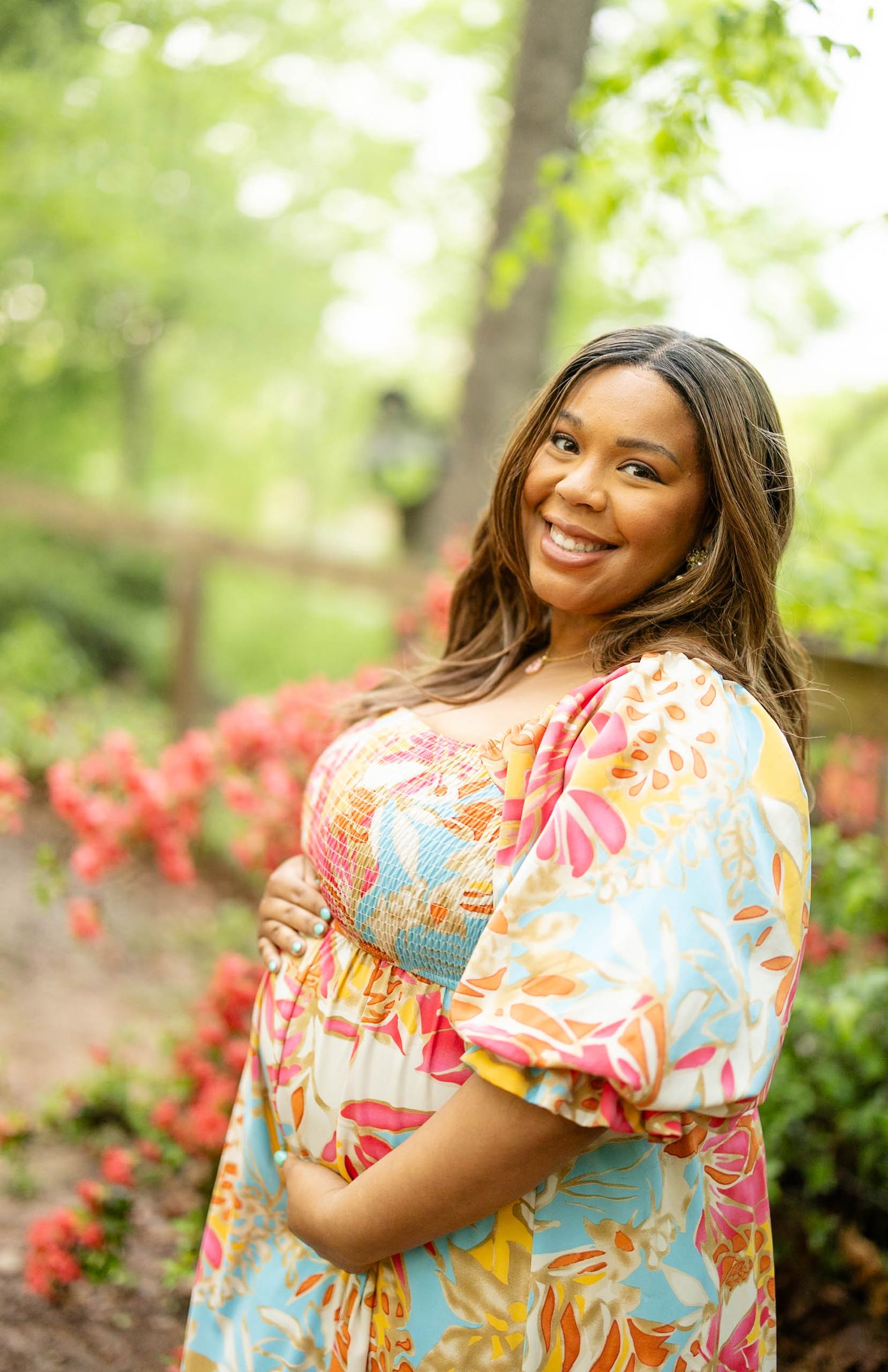Smiling pregnant woman in colorful floral dress posing outdoors among red azaleas and green trees.