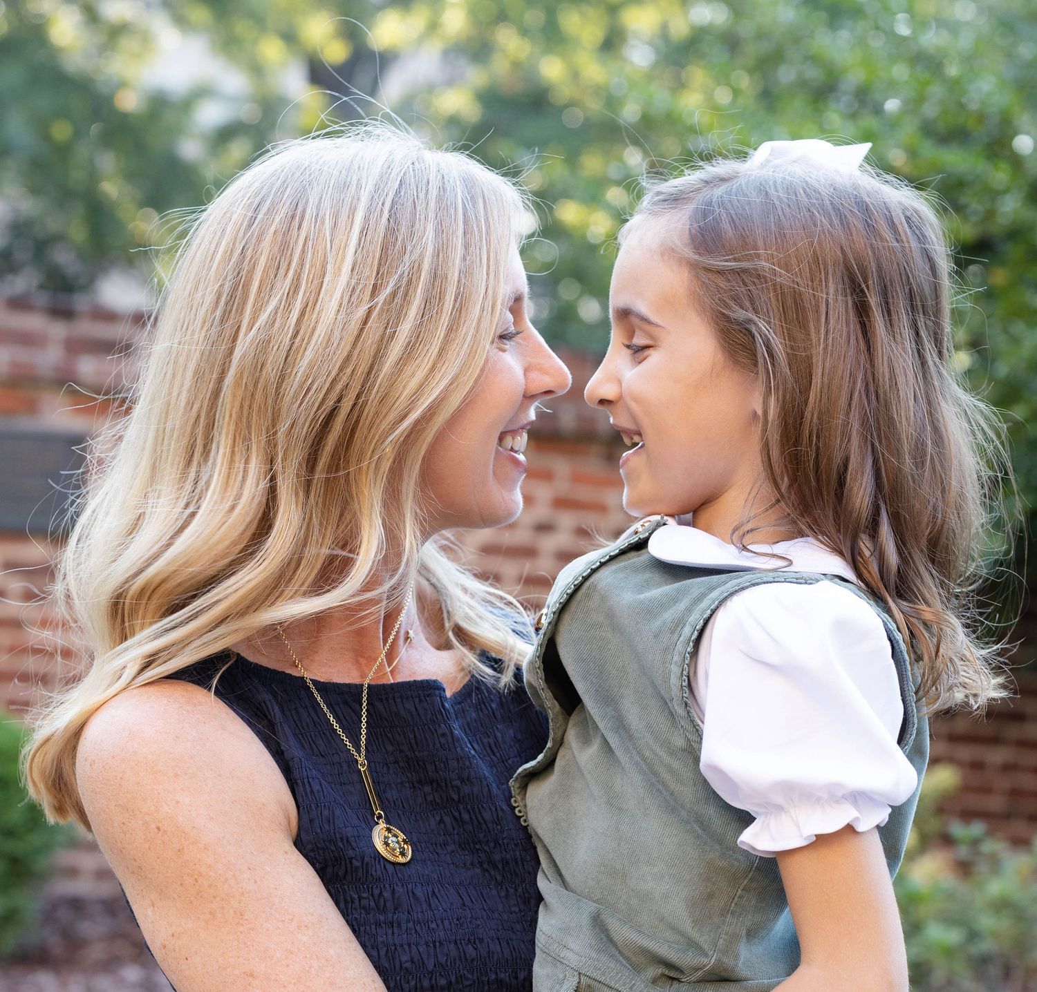 A smiling blonde woman in a navy top holds a young girl in a green vest and white blouse as they laugh together outdoors.