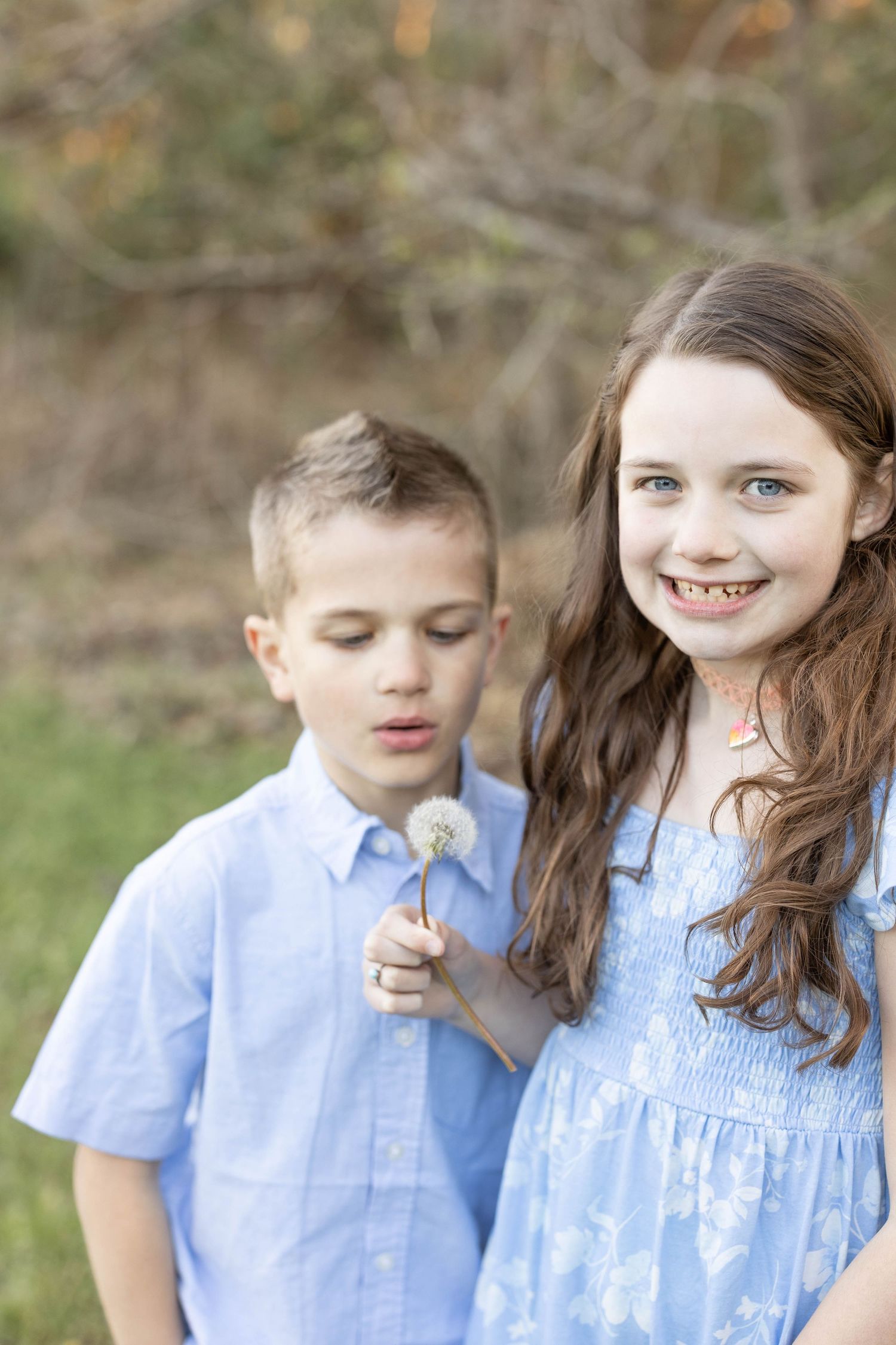 A young girl holding a dandelion while her brother blows it in a natural outdoor setting.