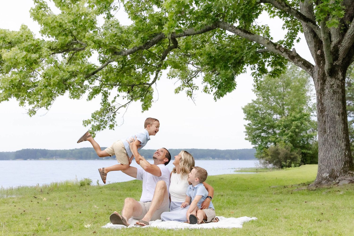 Family enjoying a sunny day outdoors by a lake, sitting on a blanket under a tree, parents playing with two young children.
