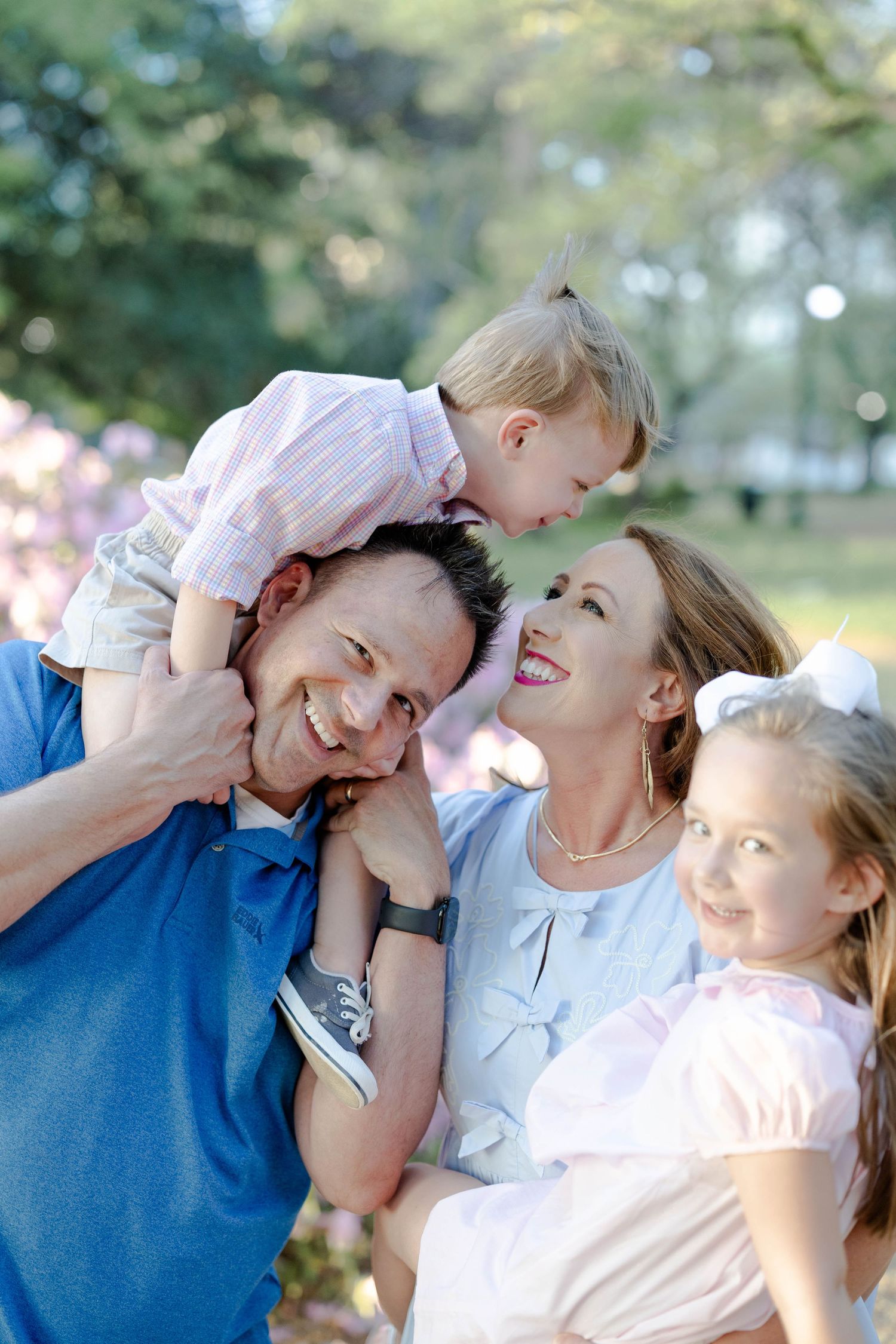 Happy family of four plays outdoors near blooming trees, father holds son on shoulders.