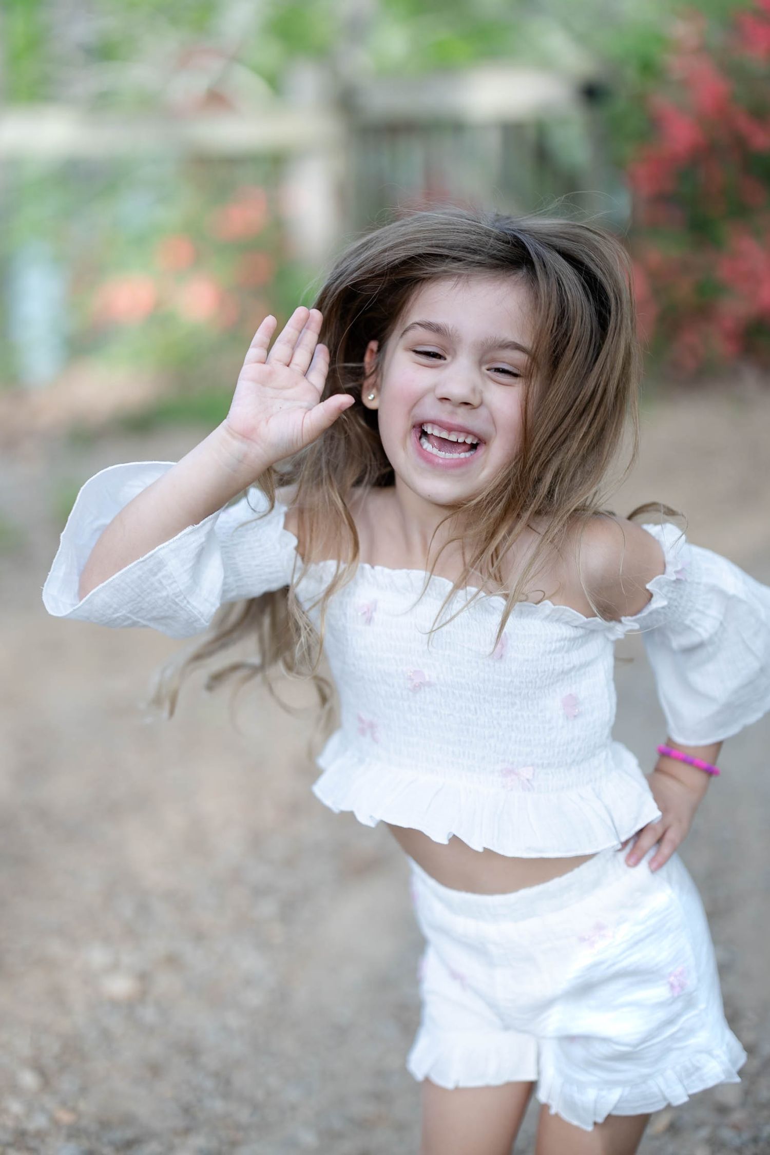 Excited young girl in white outfit laughing and waving outdoors with colorful flowers in background.