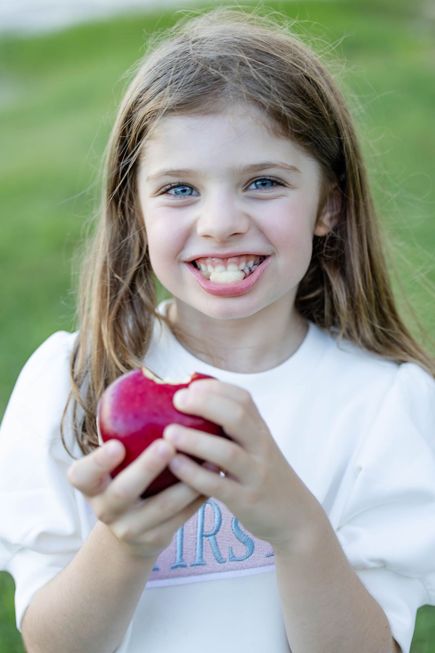 Young girl with blue eyes smiling while holding a red apple outdoors, showing missing front teeth.