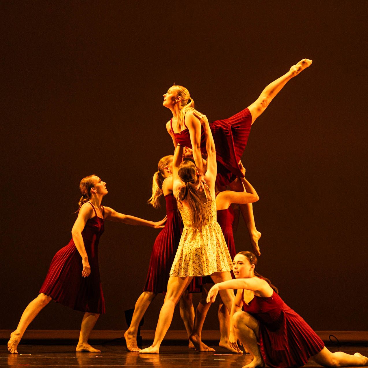 Barefoot contemporary dancers in red and yellow dresses perform a lift on a dimly lit stage as one dancer extends her leg high.