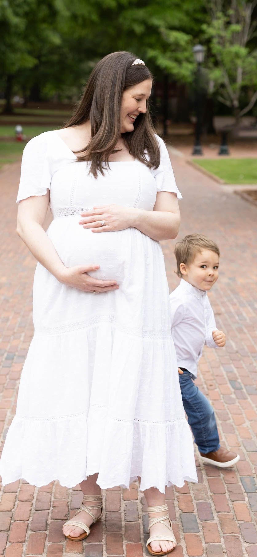 Pregnant woman in white dress holds baby bump while young toddler boy in jeans stands beside her on brick path.