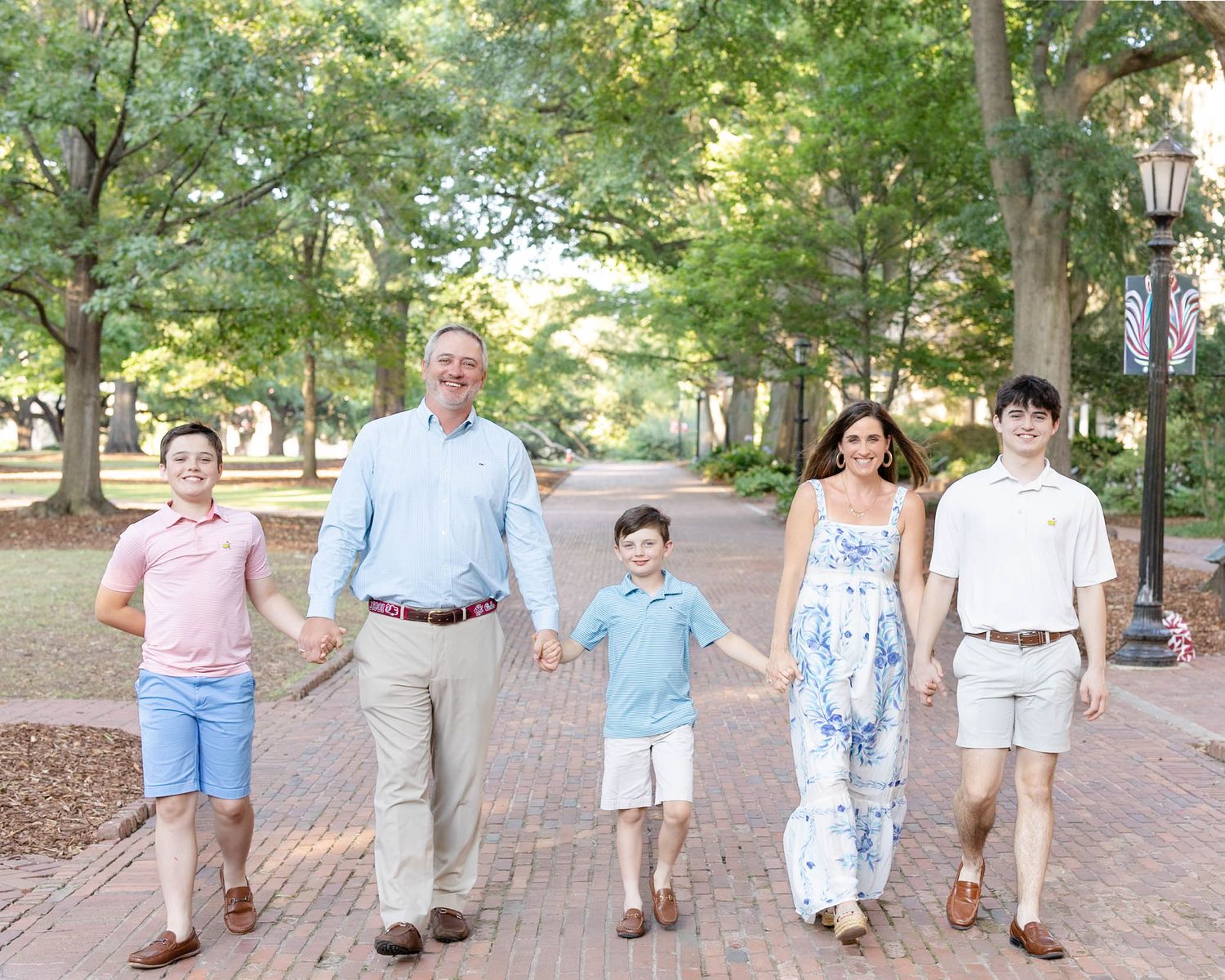 Family of five holding hands walking on brick path surrounded by lush green trees on sunny day.