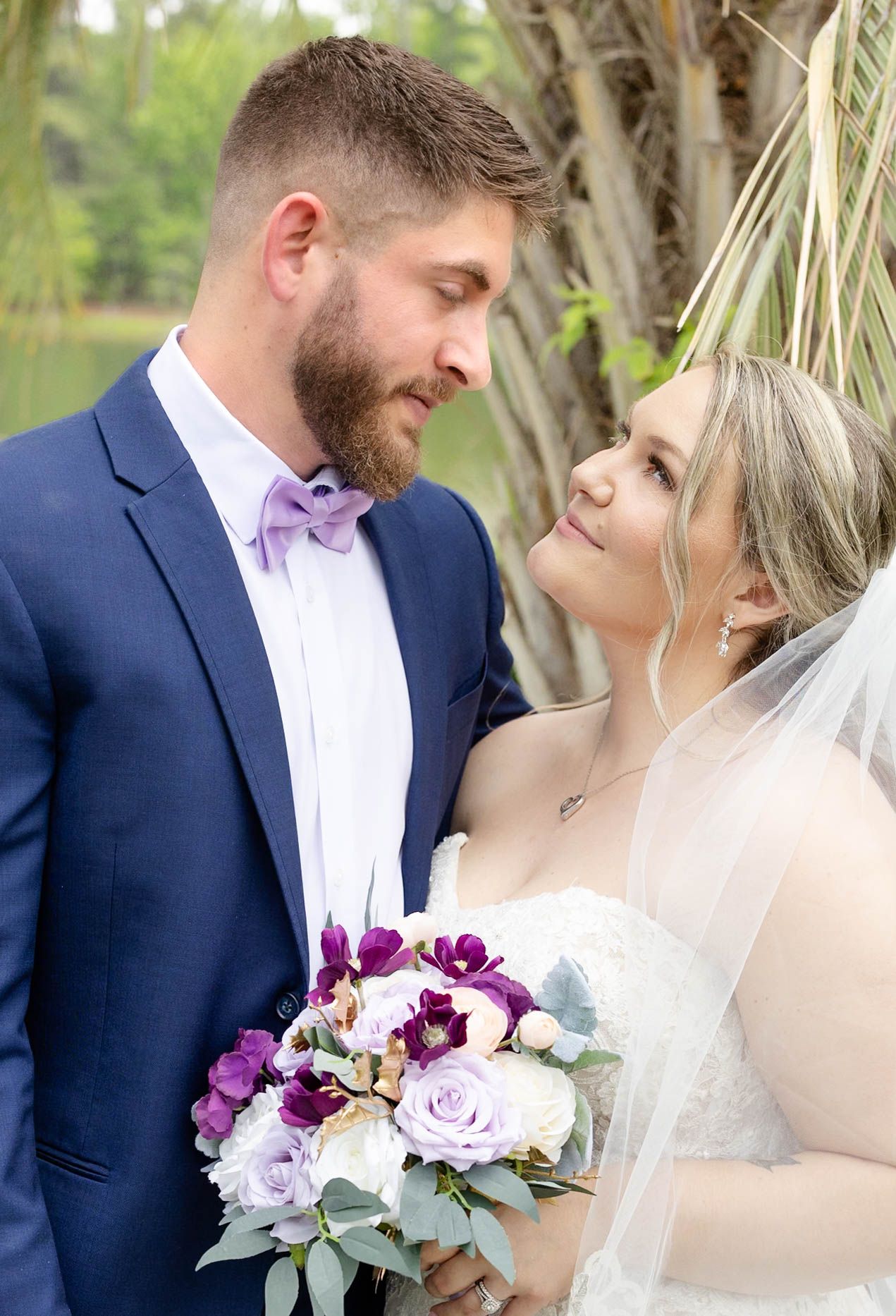 A bride and groom share a romantic moment outdoors, she holds a purple and white bouquet, he wears a navy suit with lavender bow tie.