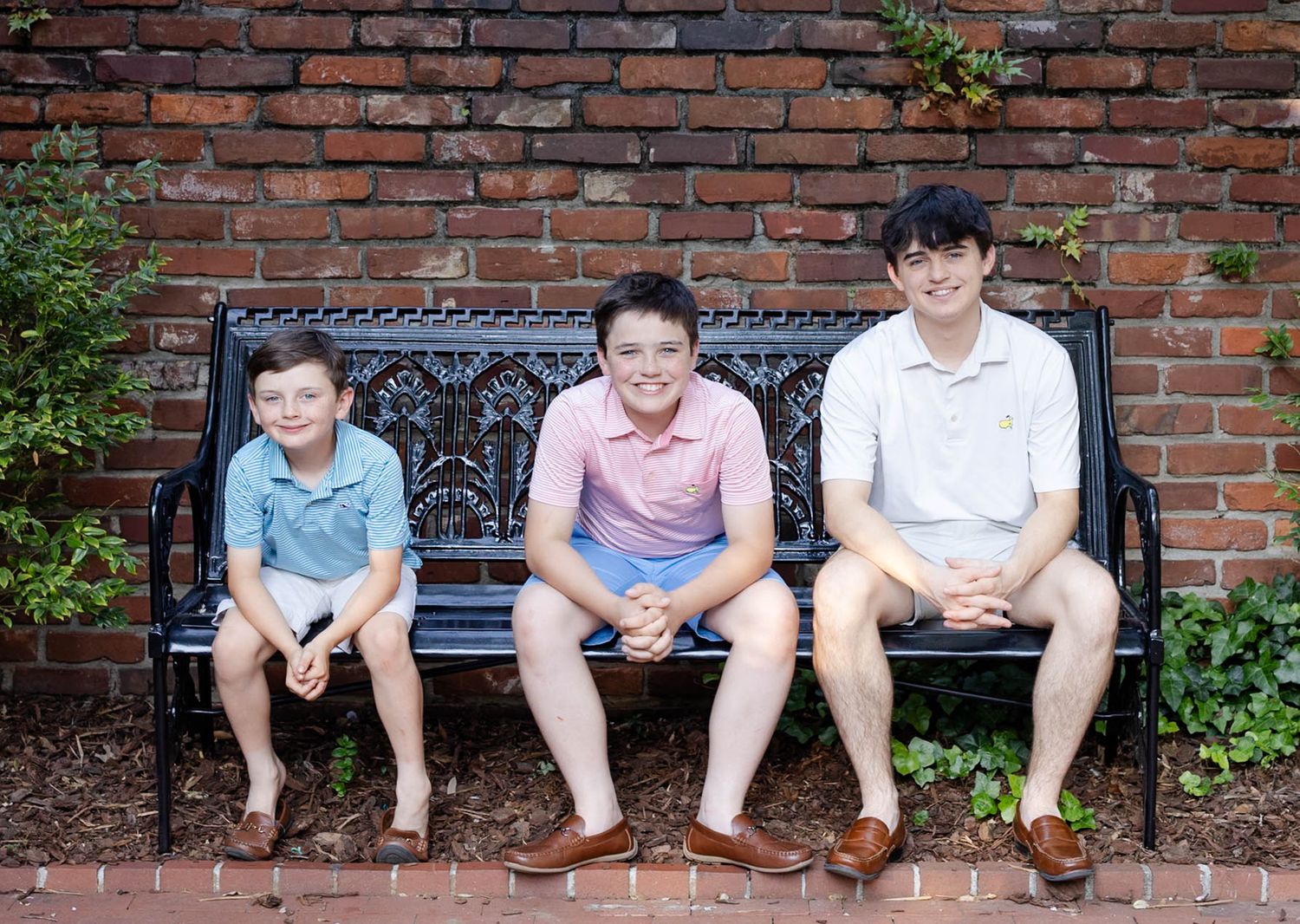 Three boys smiling and sitting together on a decorative black bench against a red brick wall outdoors.