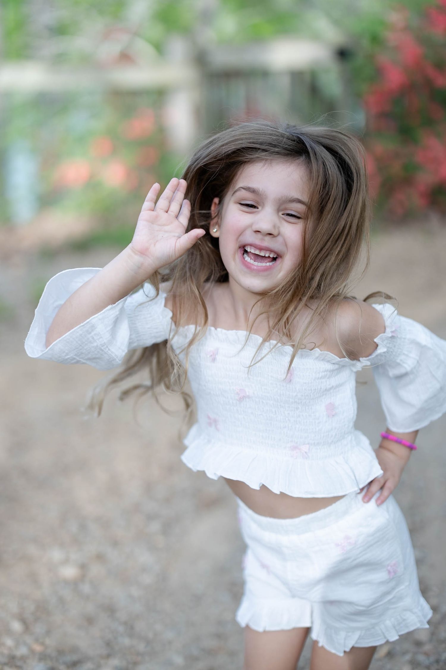 A laughing little girl in white off-shoulder top and shorts waves her hand while her hair blows in the breeze.