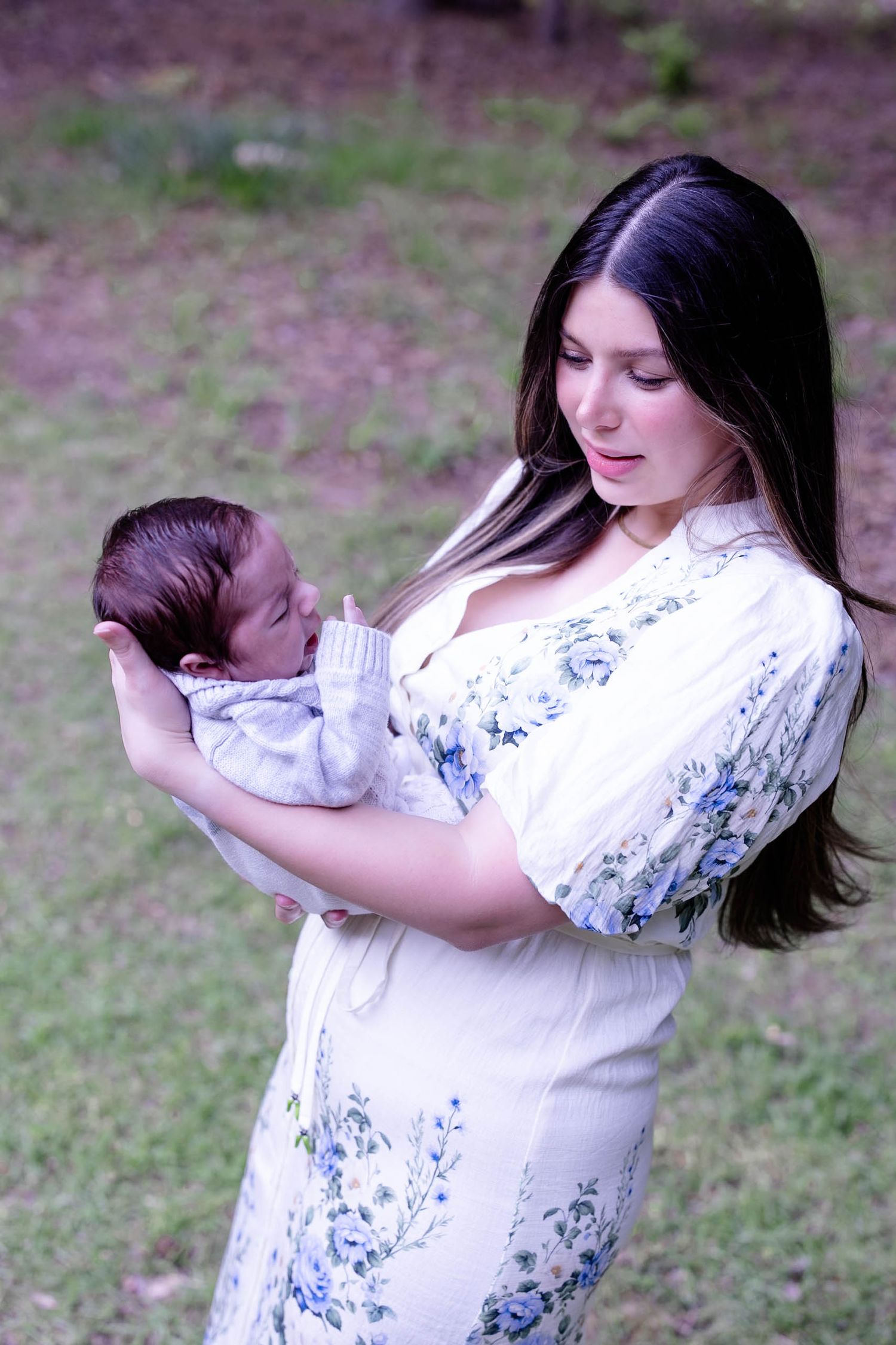 A young mother in a floral dress tenderly holds her newborn baby outdoors in a lush green setting.