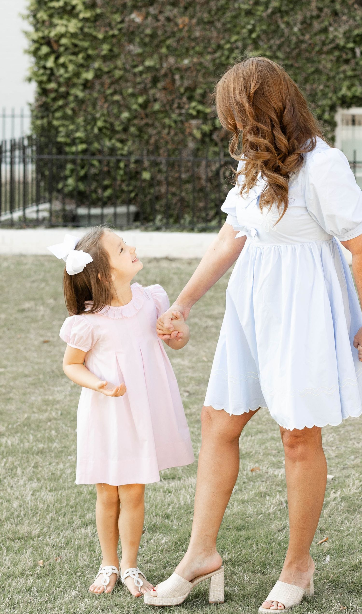 Sweet little girl in a pink dress holding hands with her mother in a light blue dress outdoors.