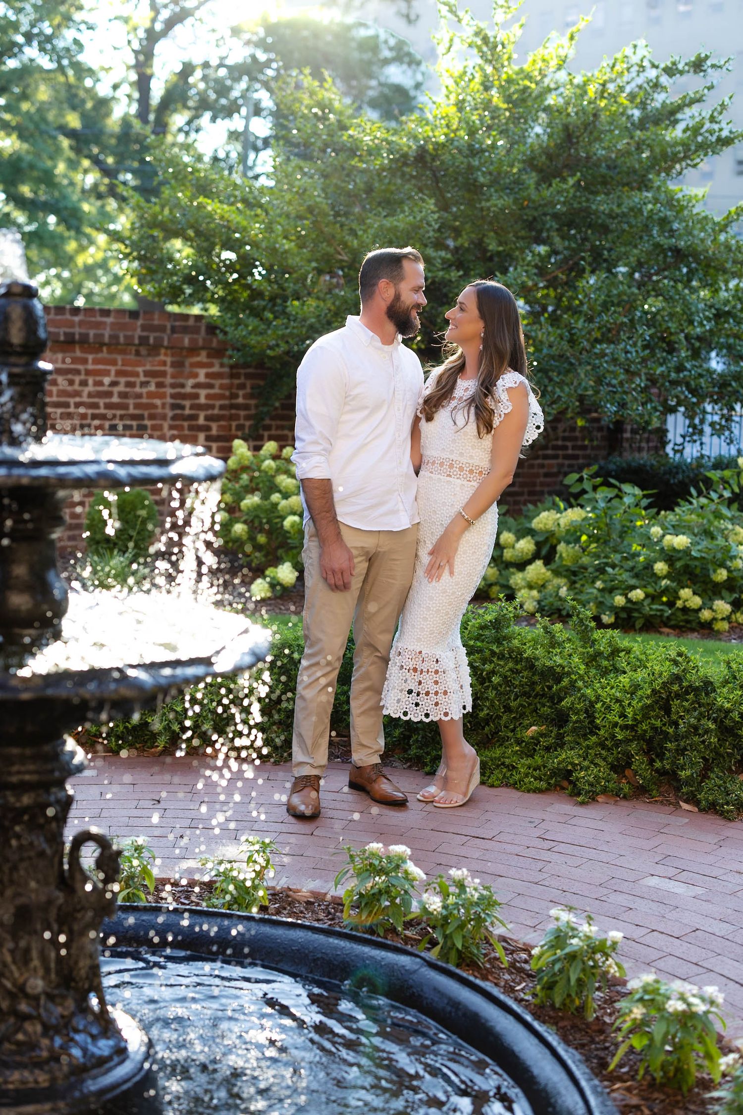 Couple sharing a romantic moment by a garden fountain, man in white shirt and khakis, woman in white lace dress.