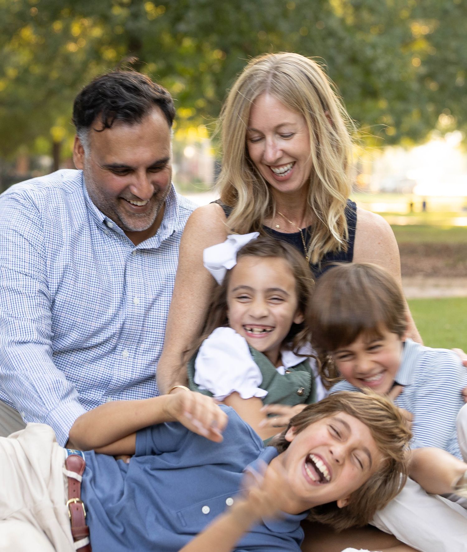A smiling family of five laughs and cuddles together on the grass in a sunny park, with three playful children.