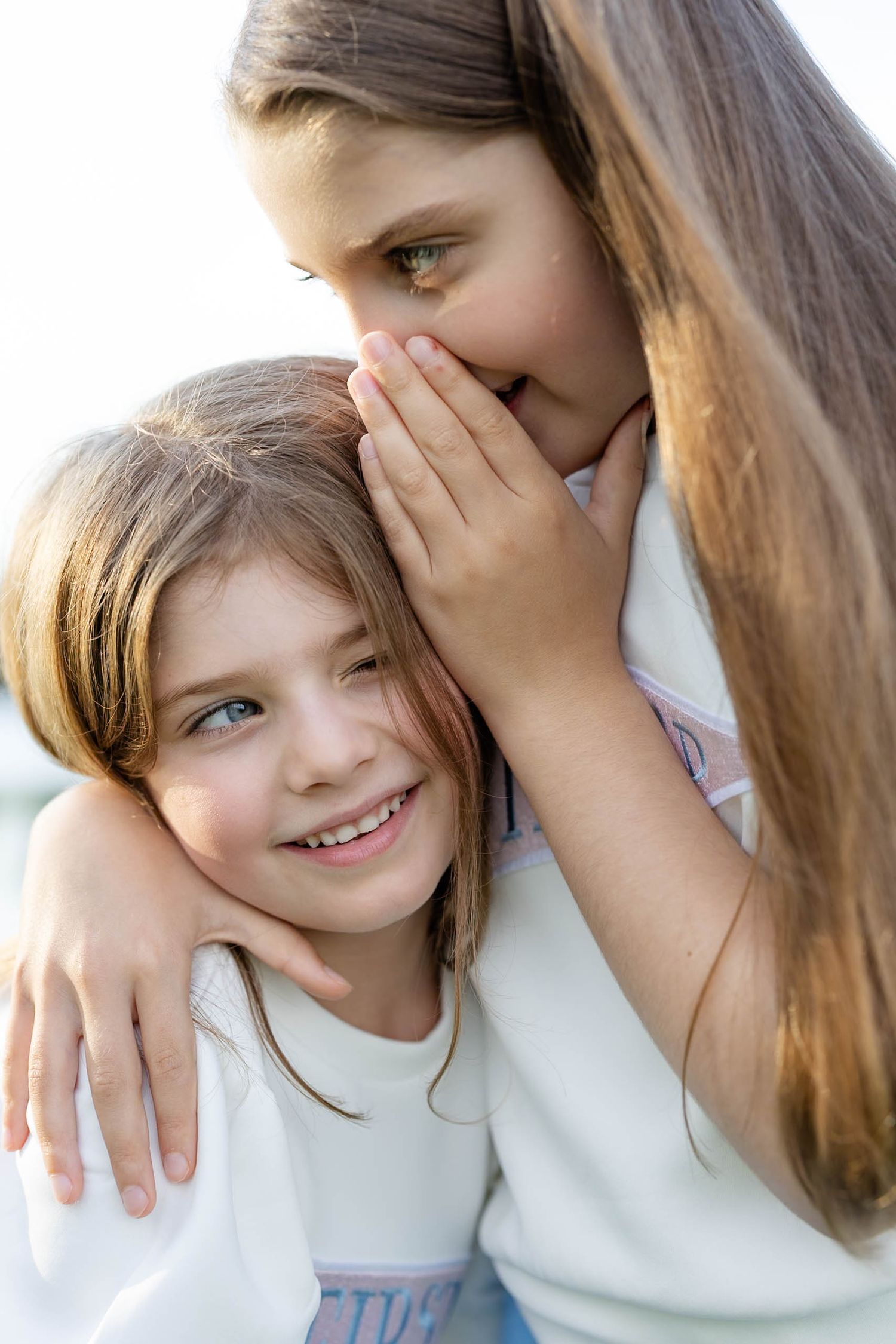 Two young girls sharing a secret whisper, one smiling brightly while the other cups her hand to her mouth.