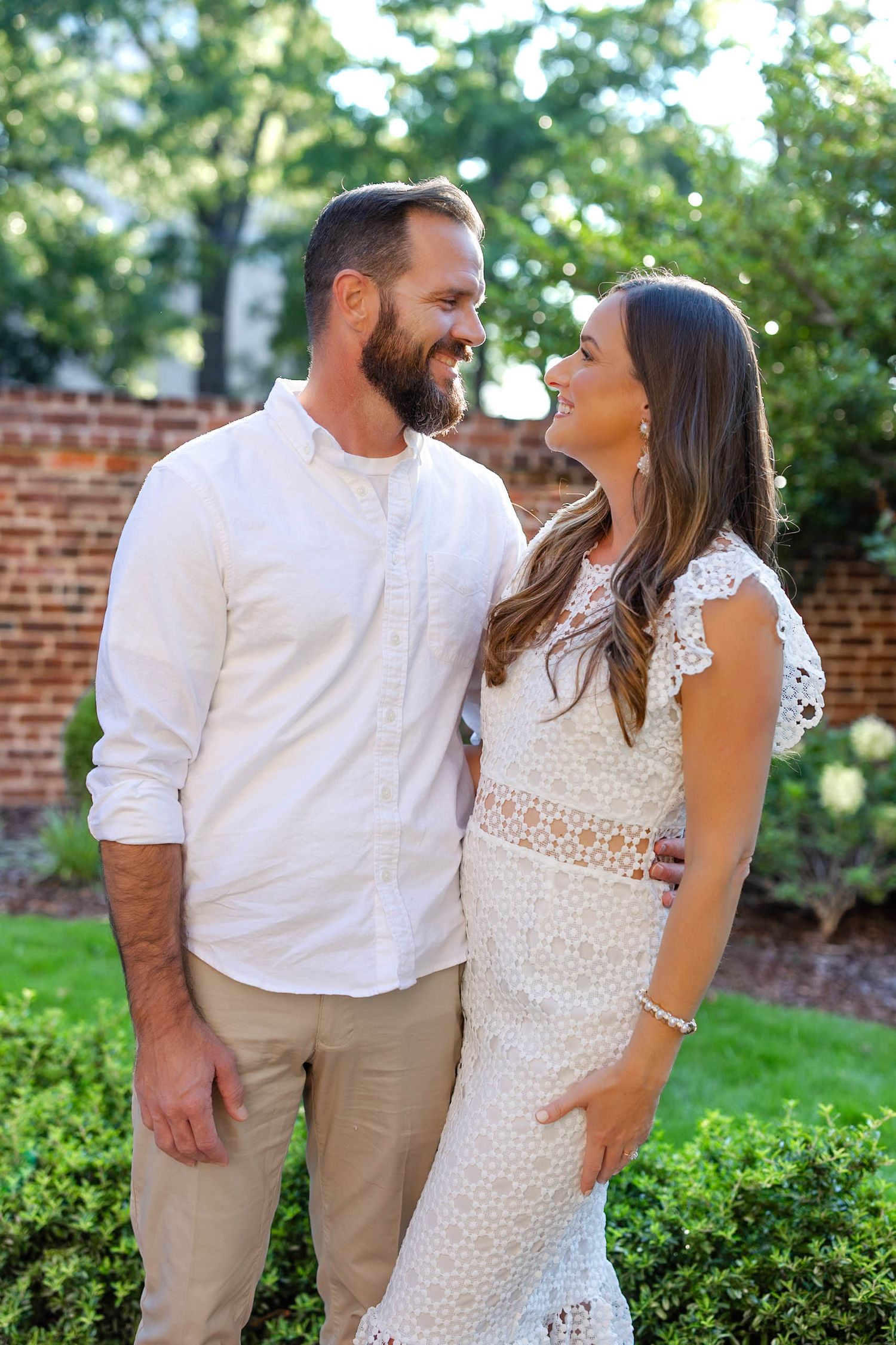 A smiling couple in coordinating white outfits stand together in a lush garden with brick wall backdrop.