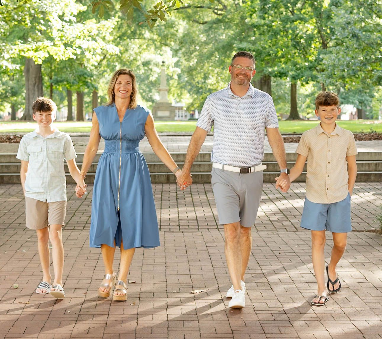 Family of four holding hands walking on brick path in sunny park with lush green trees.