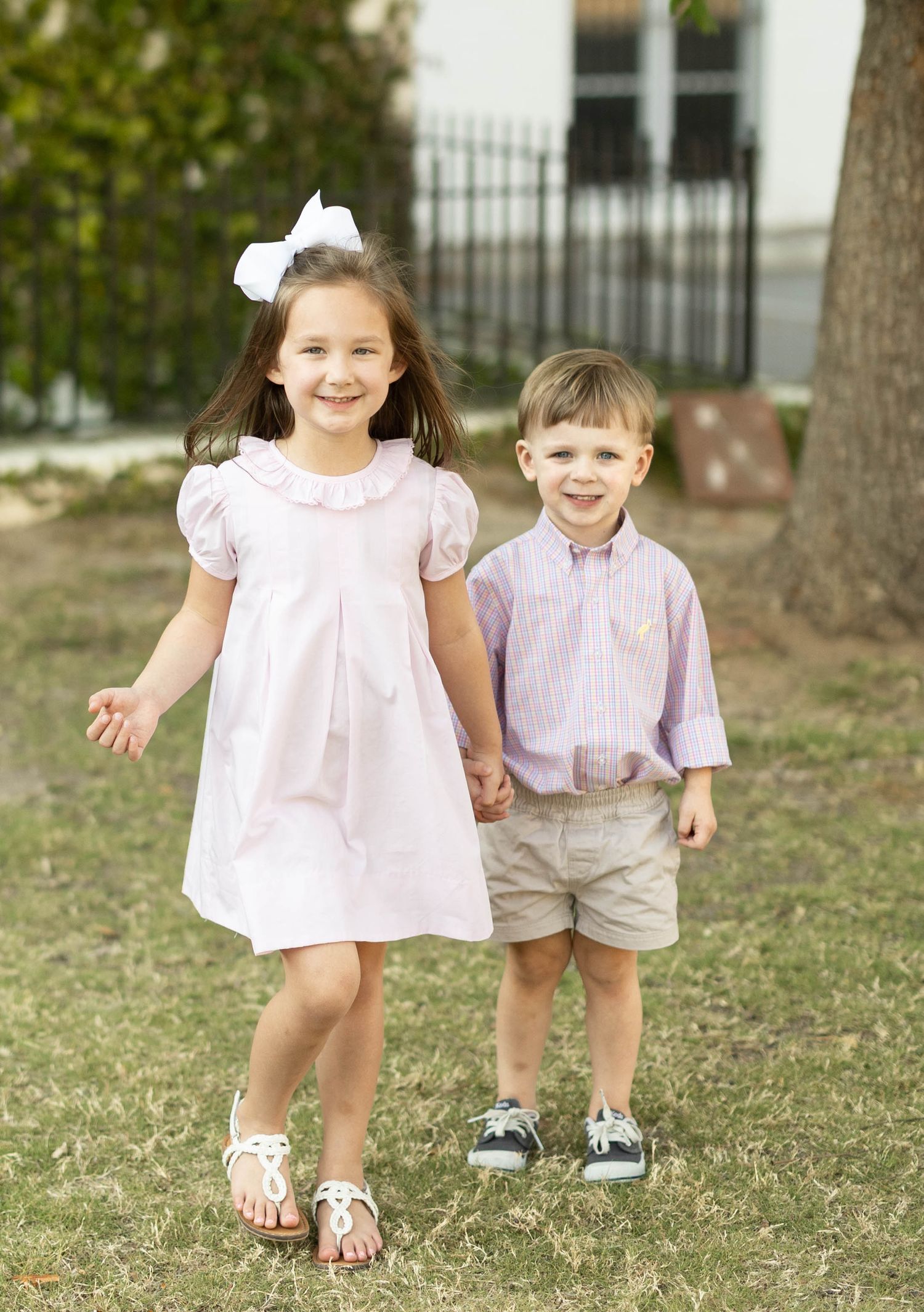 Two young siblings hold hands outdoors, girl in pink dress with white bow, boy in striped shirt.